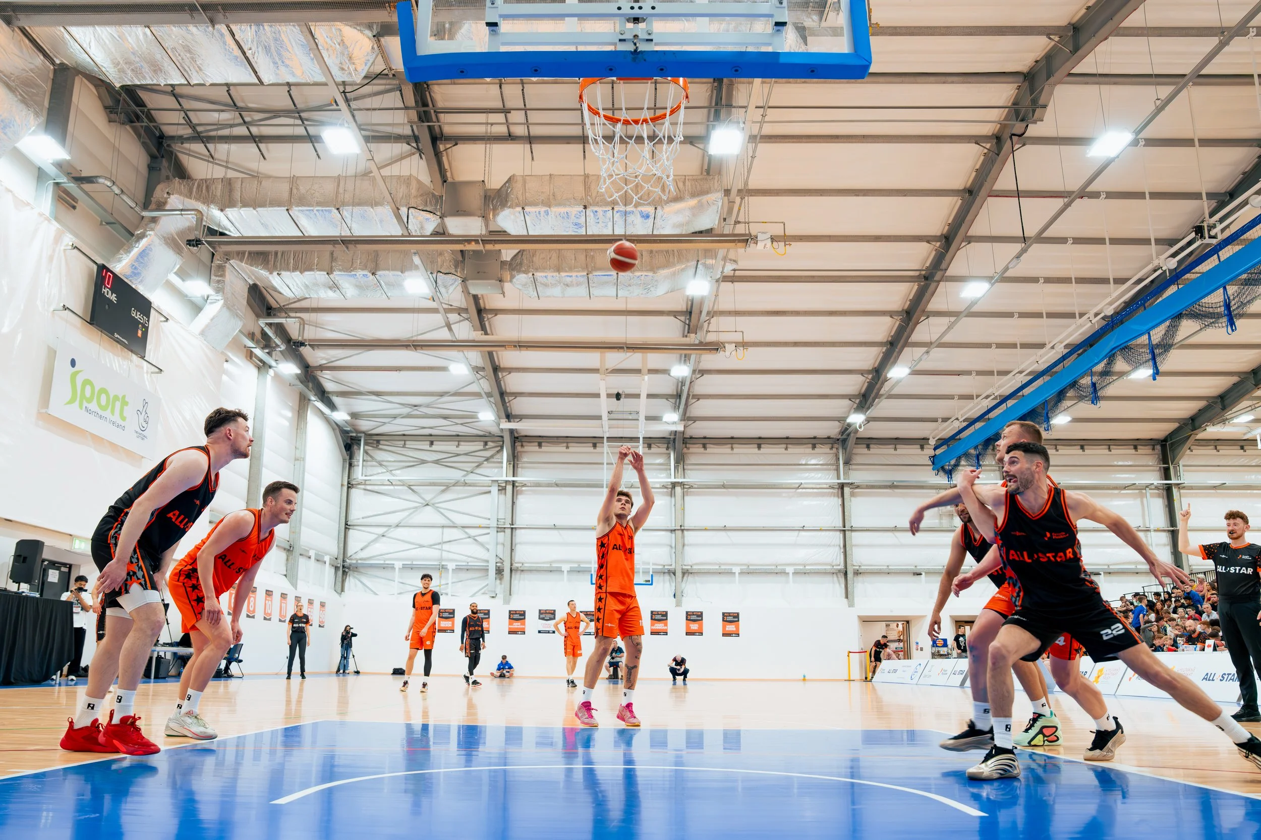 A basketball game in progress with players in black and orange uniforms on an indoor court. One player in black is about to throw the ball, while others are positioned as if preparing for rebounds or defense. Spectators are visible in the background.