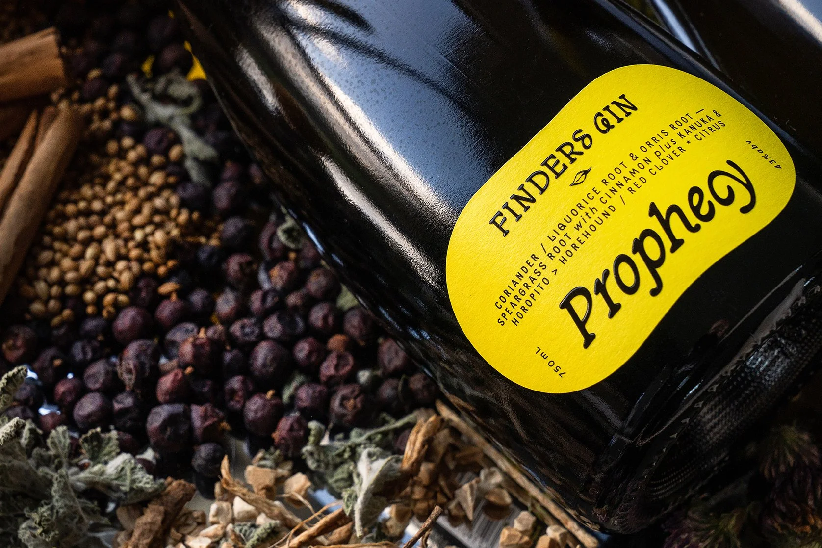 A bottle of Prophecy Fiddler's Gin lies on dried botanicals and fruit, including juniper berries, cinnamon sticks, and citrus peels.