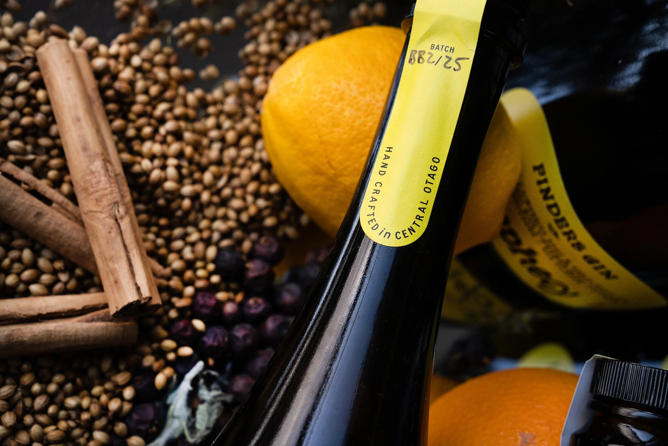 Close-up of a wine bottle with a yellow label, surrounded by oranges, cinnamon sticks, coriander seeds, black peppercorns, and a jar.