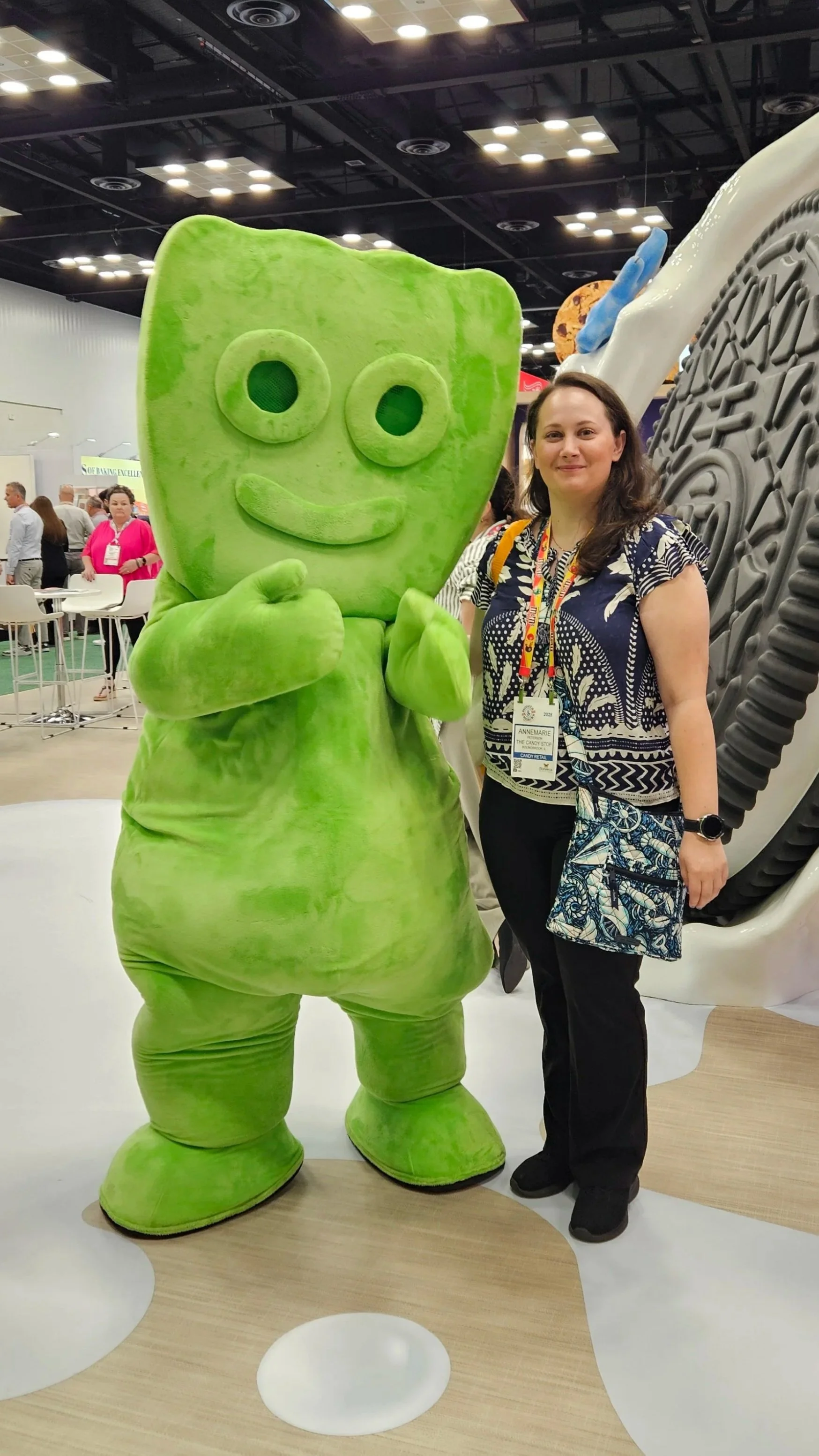A woman in a pink shirt and black pants standing next to a person in a cookie mascot costume at a trade show or convention, with colorful displays and branding related to cookies and milk in the background.