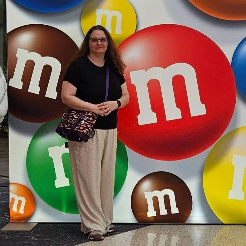 A woman in a red shirt holding a shopping bag, standing in front of a backdrop with large colorful M&M's logos.