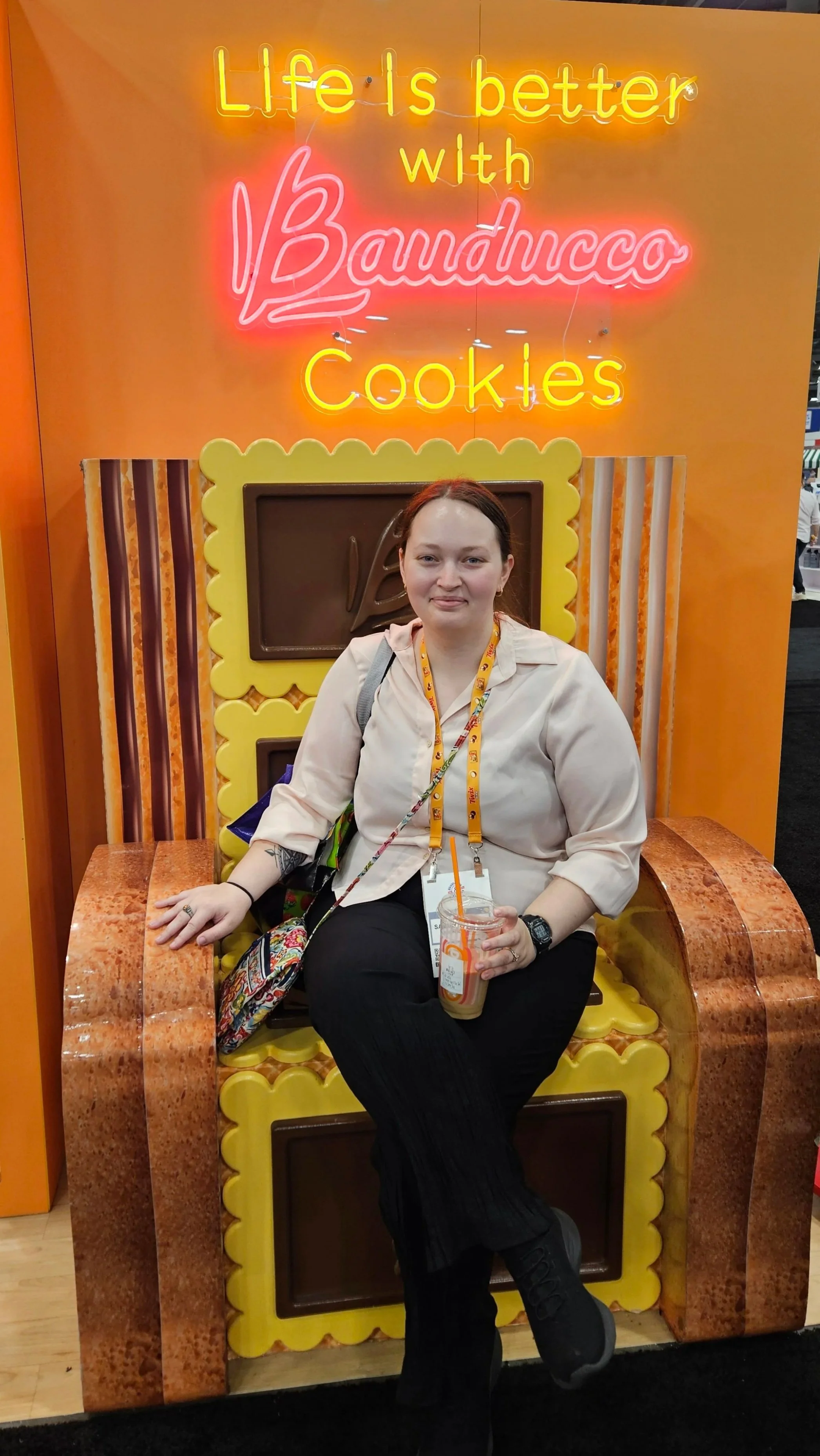 A young woman sitting on a large, ornate throne with a high backrest adorned with vintage-style illustrations. She wears a T-shirt, shorts, sunglasses on her head, and has a camera around her neck. She is in an arcade or gaming store, surrounded by shelves filled with collectible items.