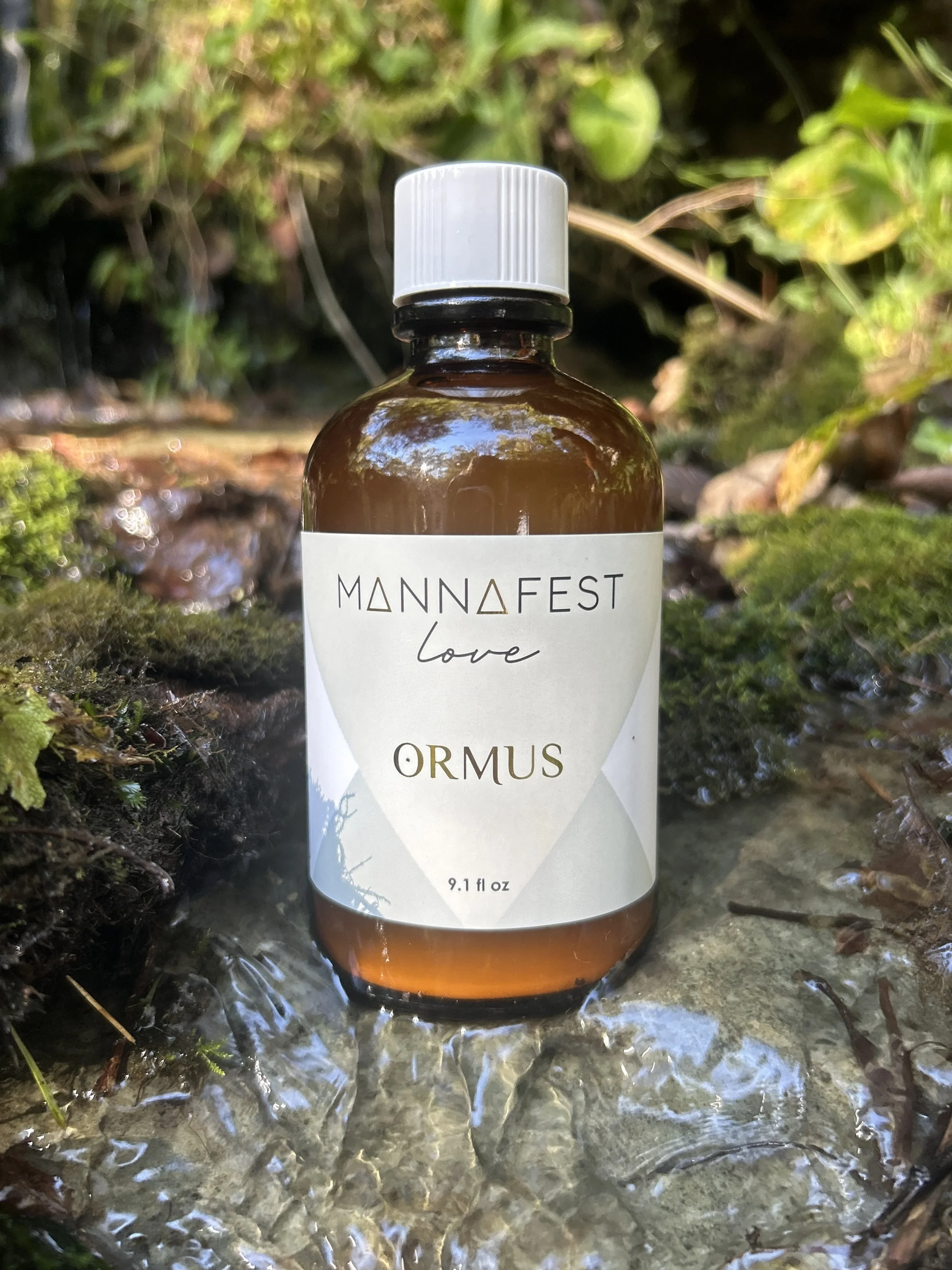 A brown glass bottle of Ormus with a white cap and label, placed on a shallow stream with moss and green plants around it.