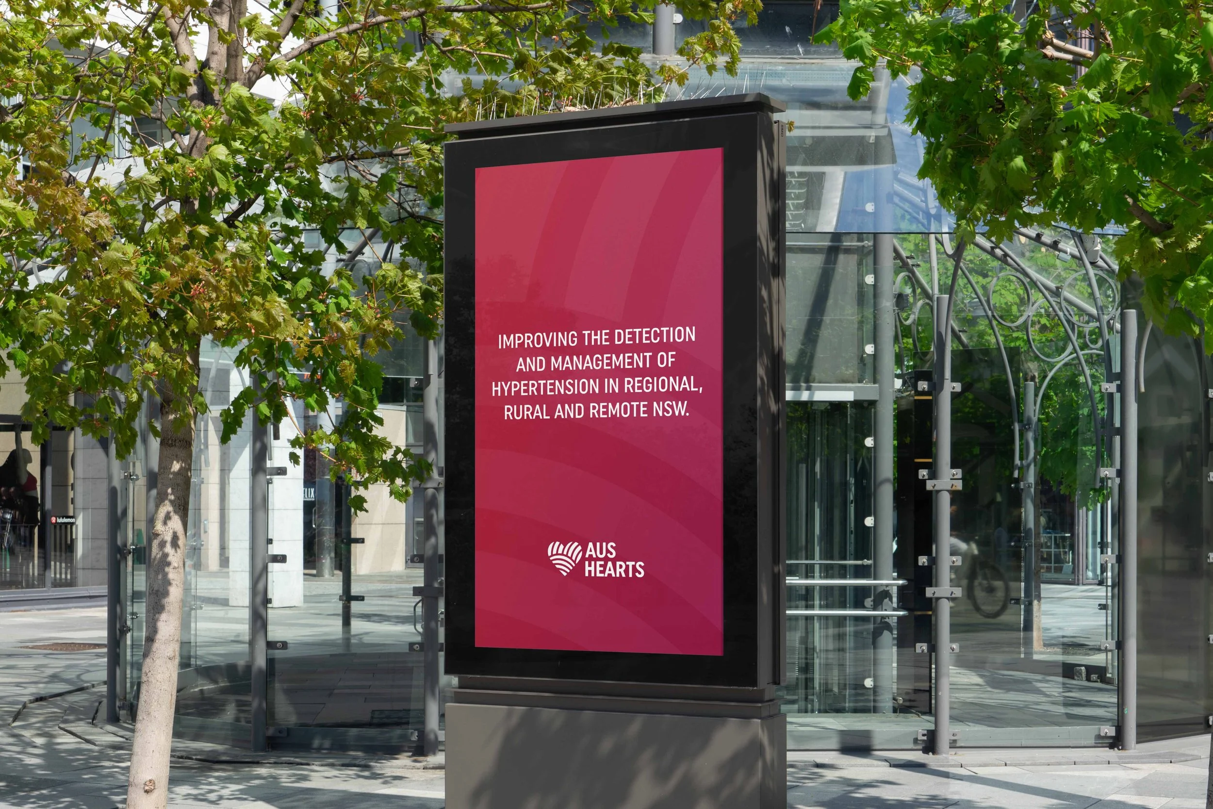 A digital billboard displays a message about improving detection and management of hypertension in regional, rural, and remote NSW, with the AUS HEARS logo at the bottom. The billboard is located outdoors near trees and a glass bus shelter.