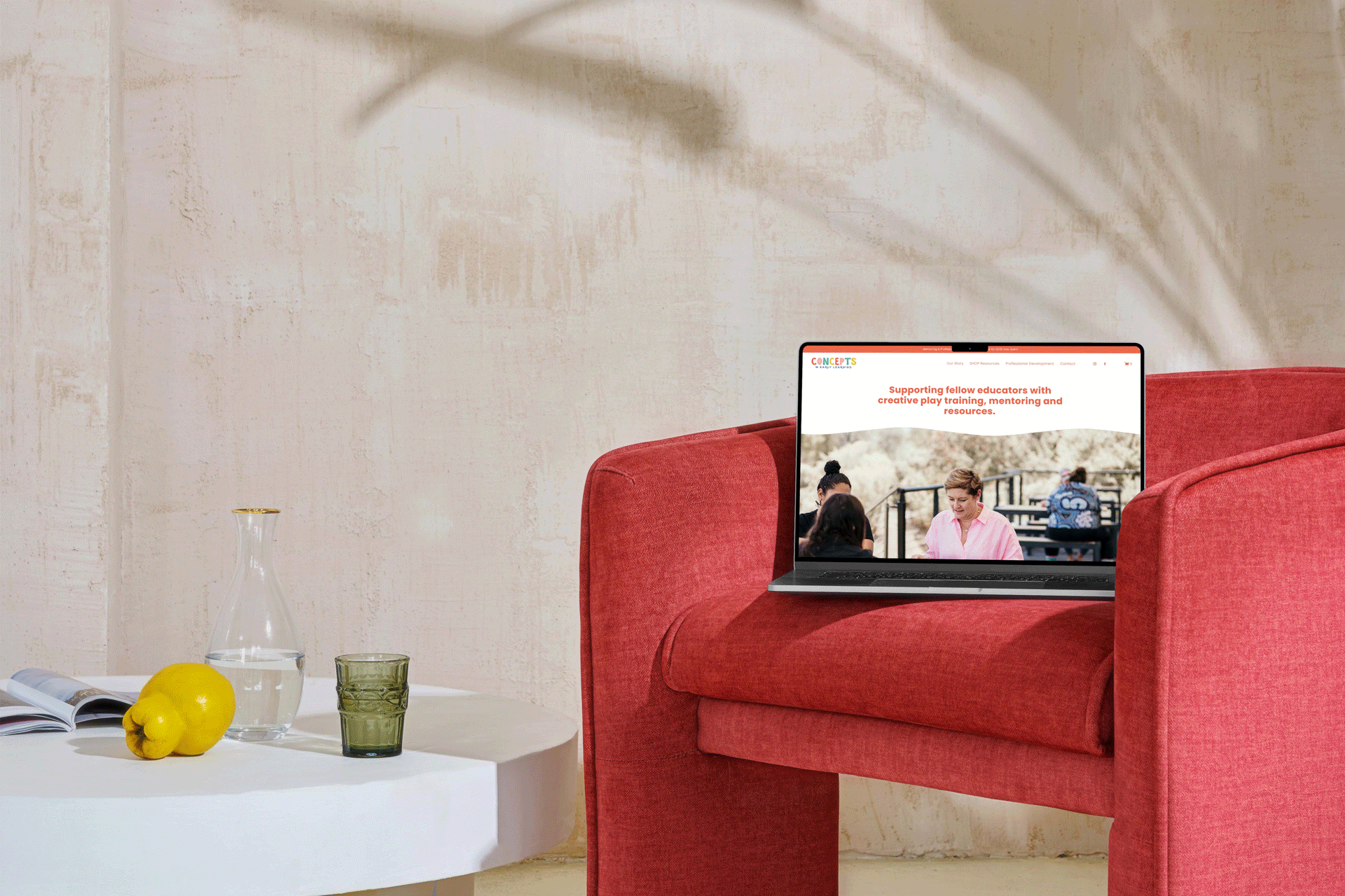 A red armchair with a laptop displaying a website about supporting educators sits on the floor. A white side table next to it holds a clear glass pitcher, a yellow bell pepper, a drinking glass, and an open magazine. The background is a textured light-colored wall.