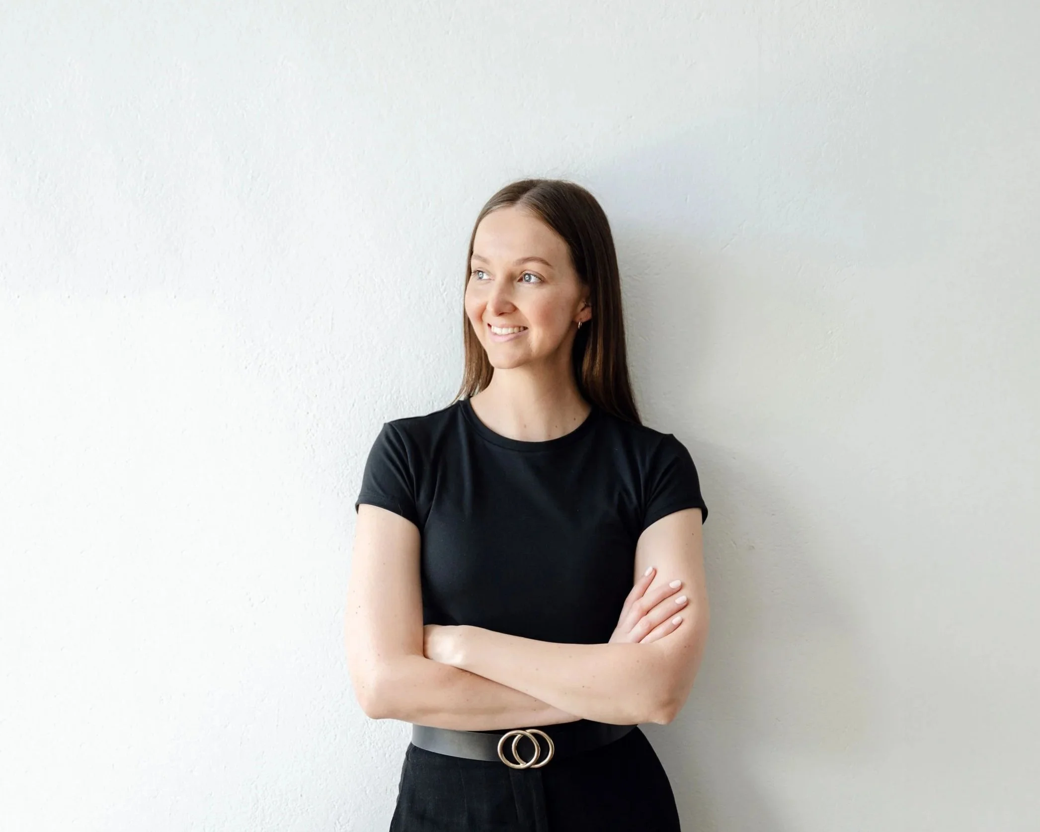 A young woman with long brown hair, wearing a black t-shirt and black skirt with a double-ring belt, standing with her arms crossed and smiling while looking to her left against a plain white wall.