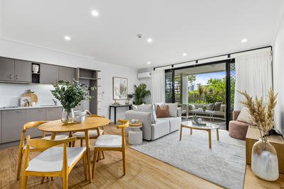 Modern open-concept living and dining area with wooden furniture, neutral decor, and a view of a garden through large sliding glass doors. The kitchen is compact with gray cabinets, and the space is well-lit with natural and recessed lighting.