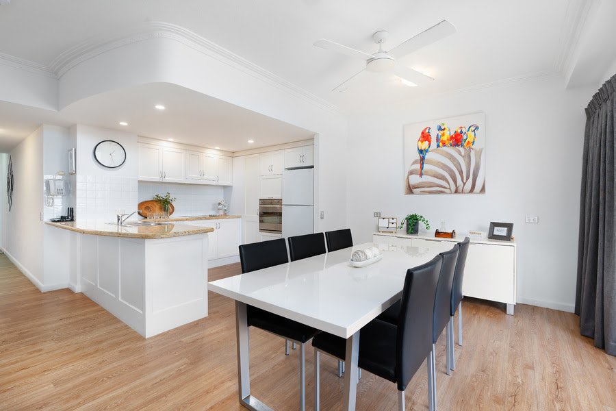 Modern kitchen and dining area with wooden flooring, white cabinets, a clock on the wall, a dining table with black chairs, and a colorful painting of birds hanging on the wall.