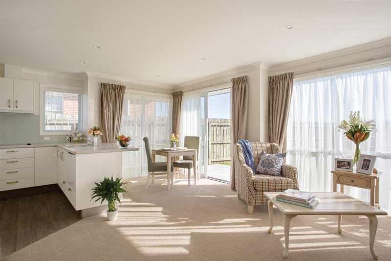 Bright living room with a plaid armchair, side table, small dining area, potted plants, and large windows with curtains.