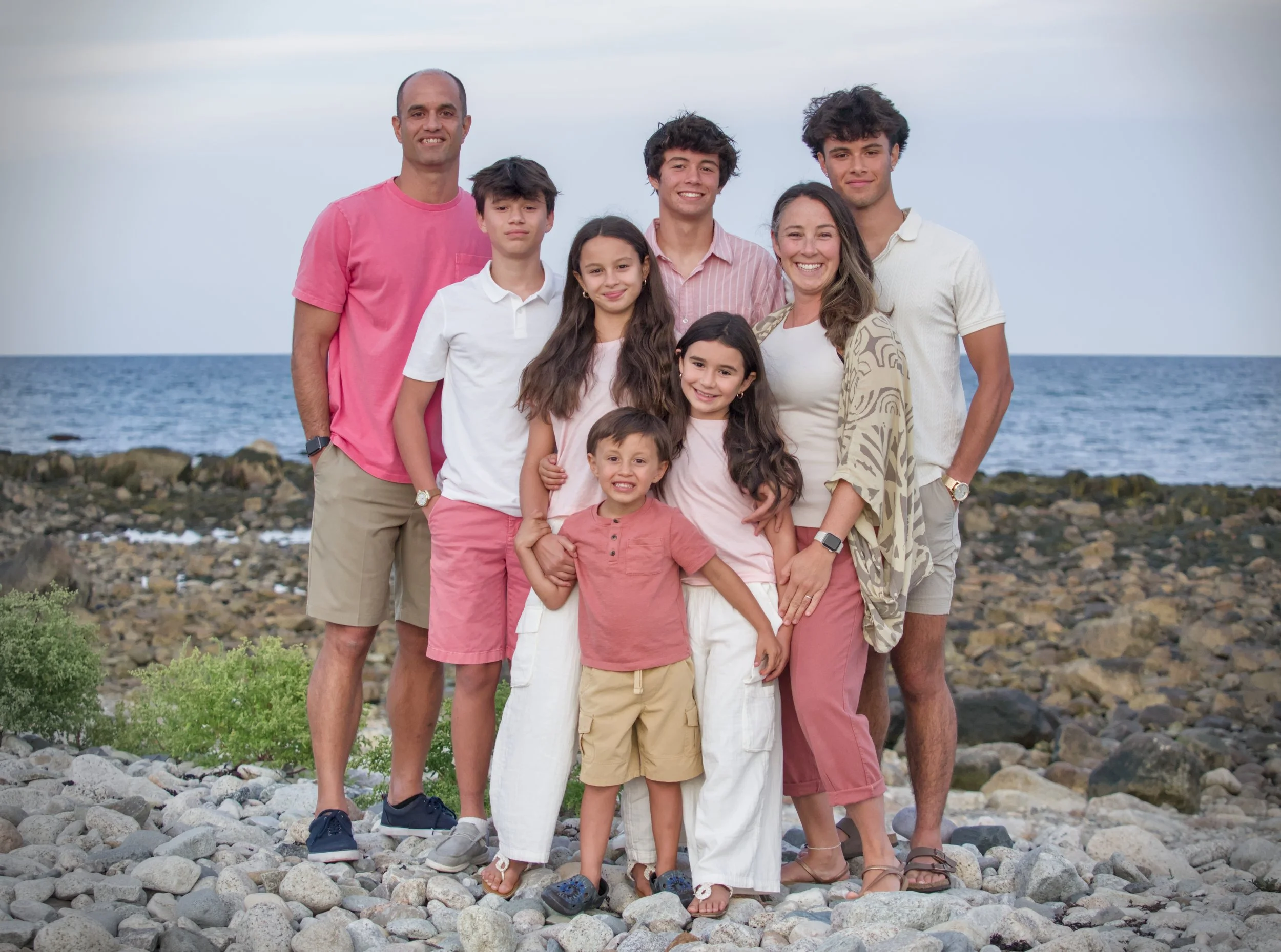 Family standing on rocky beach with ocean and sky in the background, all smiling warmly.