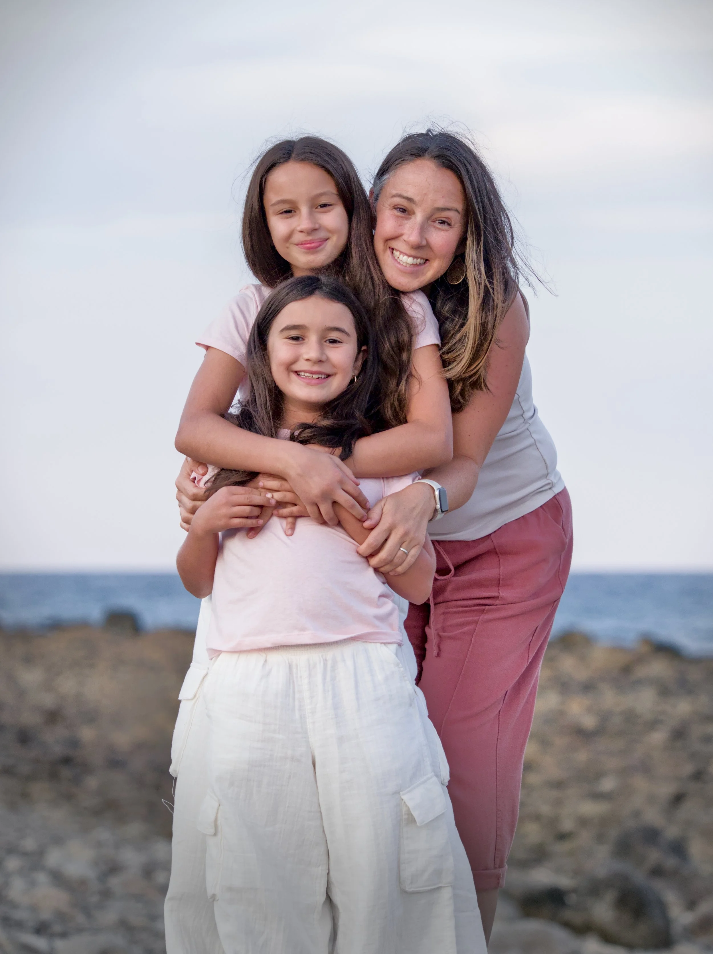 A woman and two young girls smiling on a rocky beach during sunset, with the ocean in the background.