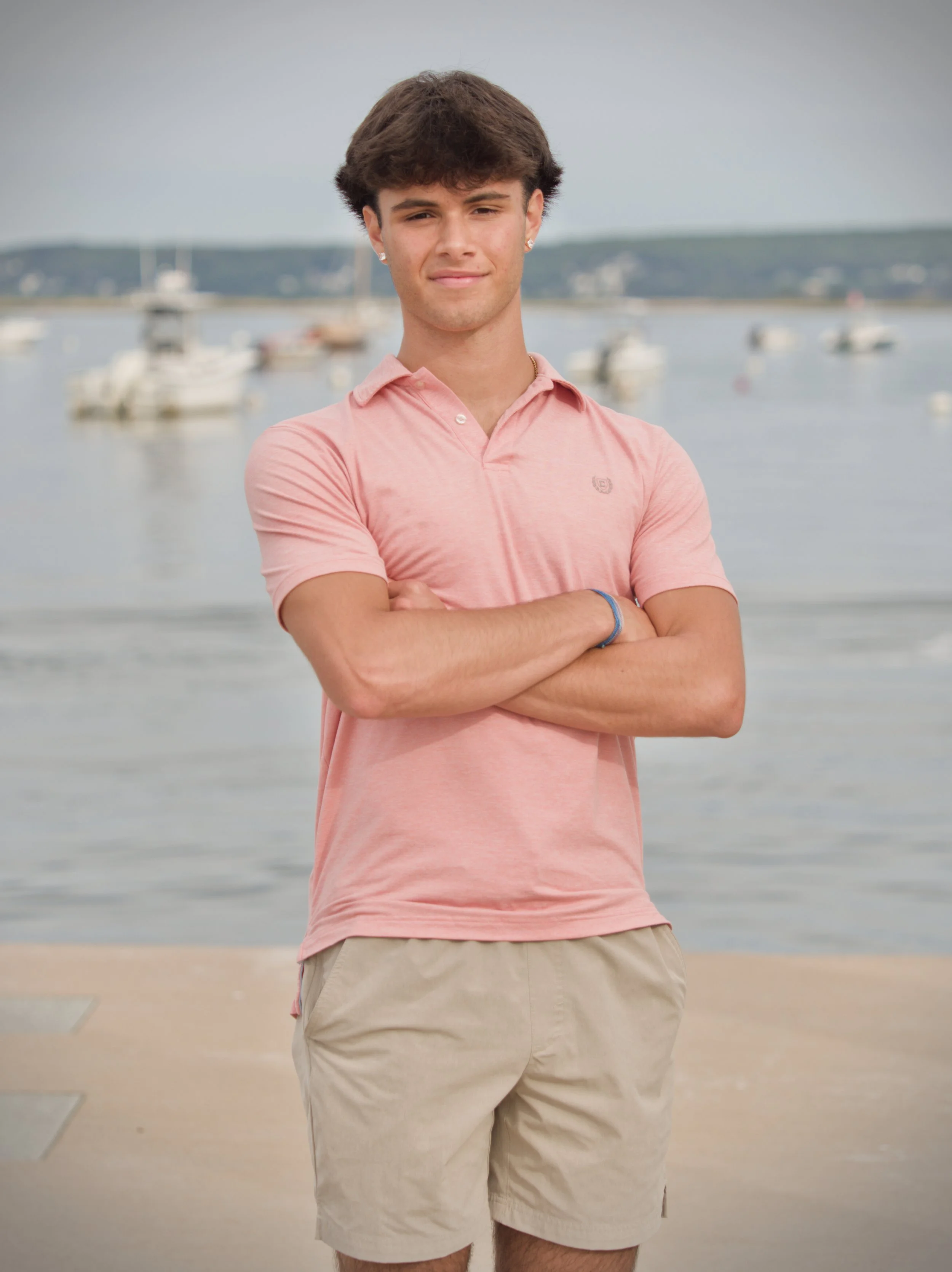 A young man with brown hair, wearing a pink polo shirt and beige shorts, standing with arms crossed in front of a body of water with boats in the background.