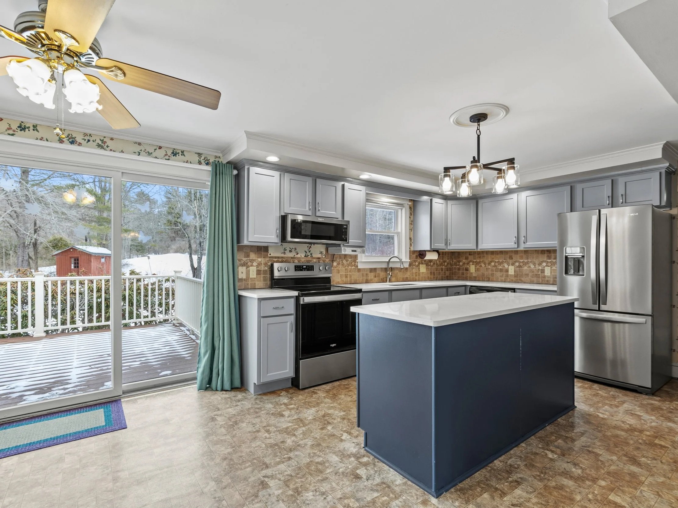 Modern kitchen with gray cabinets, stainless steel refrigerator, black stove, microwave, and a white island with a blue base. There is a sliding glass door leading to a deck outside, which has snow and trees.