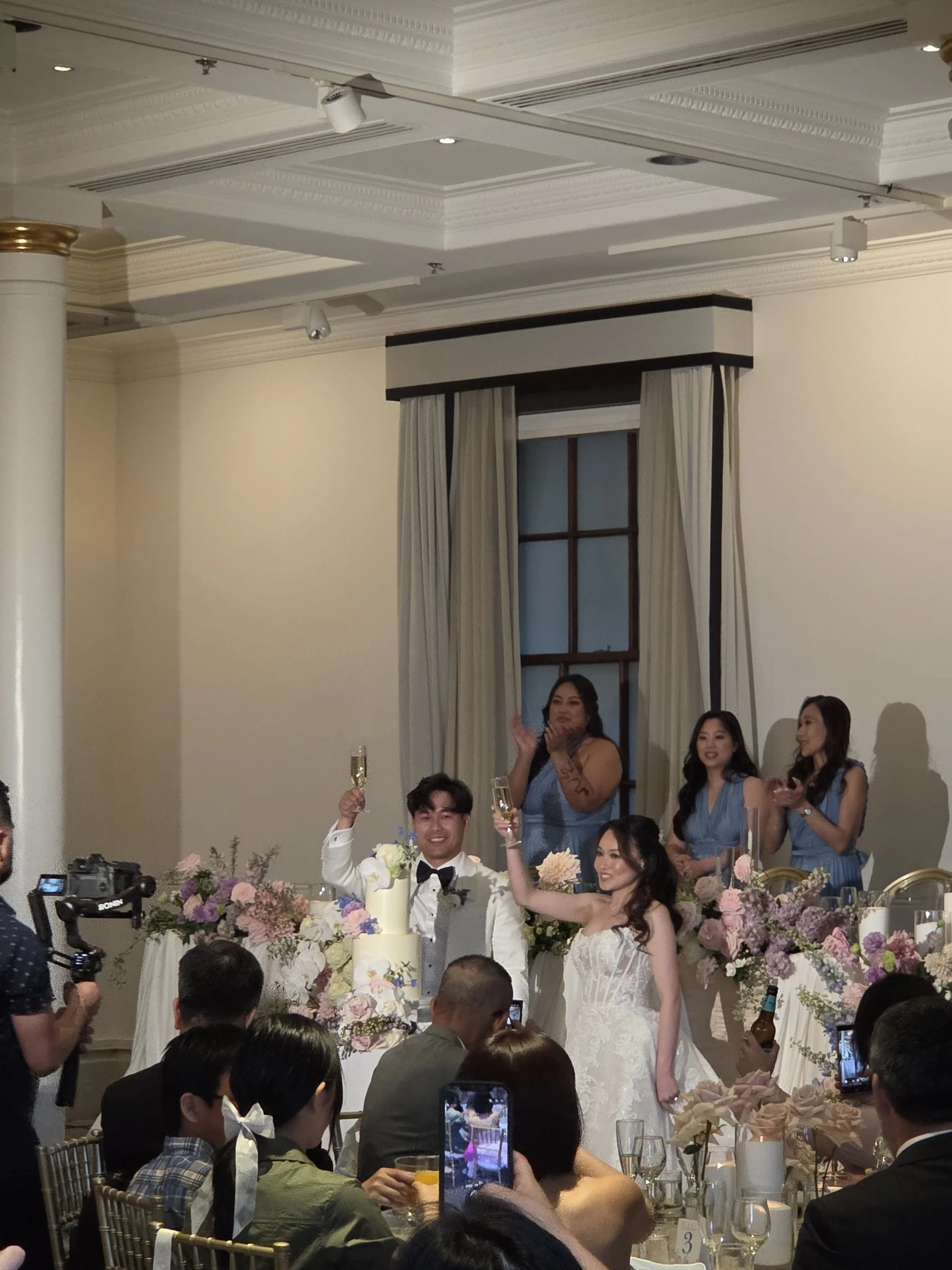 A wedding celebration with the bride and groom raising glasses of champagne, surrounded by friends and family in an elegant room with floral decorations.