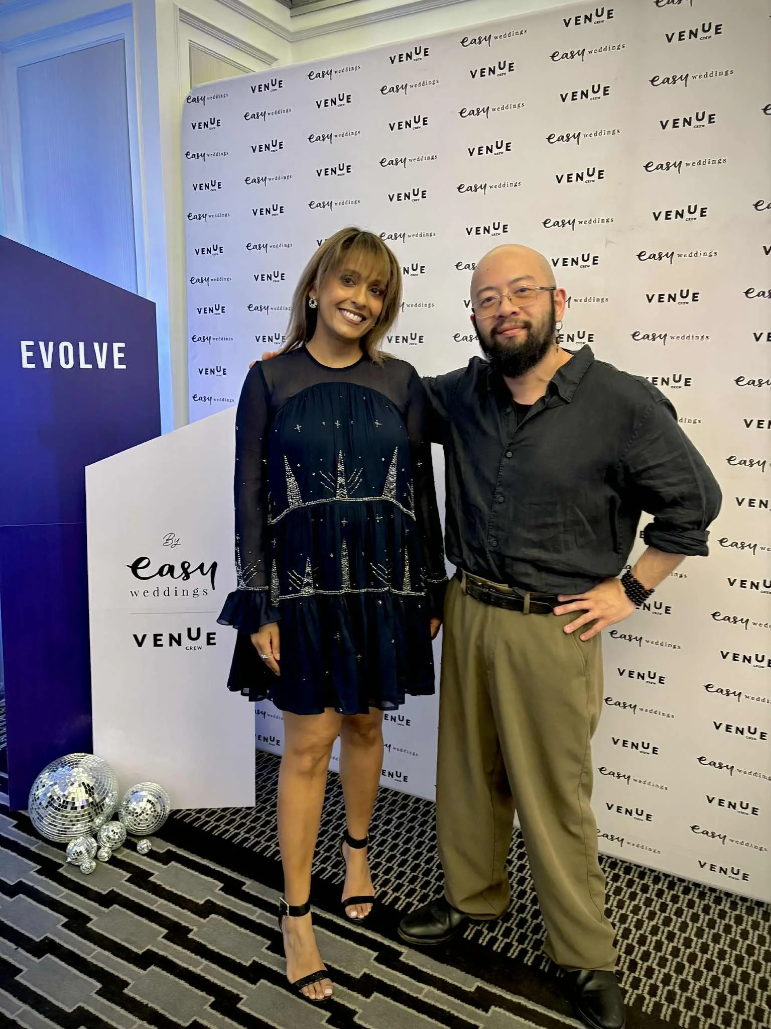 A woman in a black dress and heels and a man in a black shirt and khaki pants stand together at an event, smiling. The backdrop features logos for easy weddings and venue, and there are disco balls on the floor nearby.