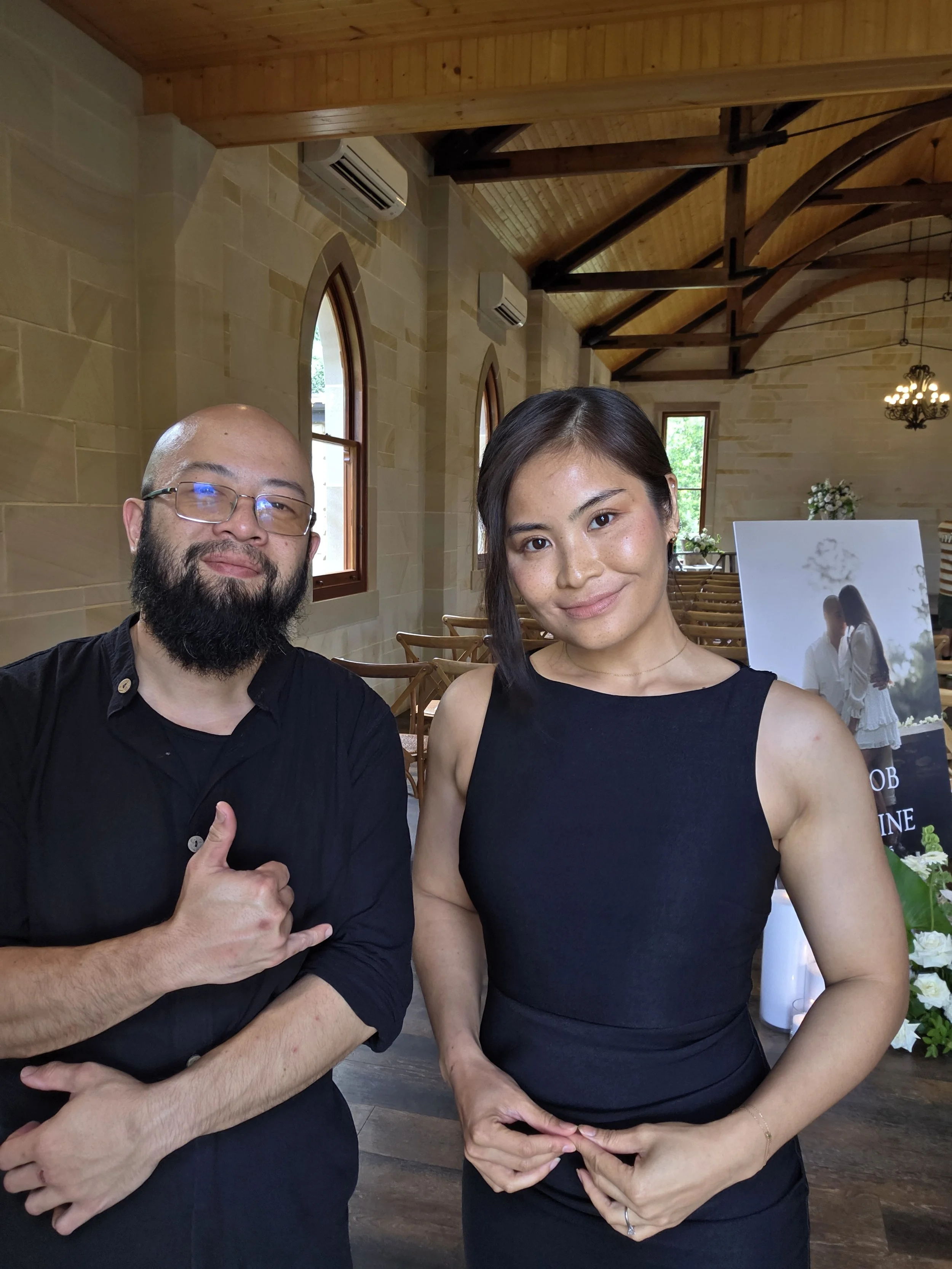 Two people posing for a photo inside a church, with wooden ceiling beams and arched windows in the background. One is a bald man with a beard and glasses, wearing a black shirt, making a hand gesture resembling a sign. The other is a woman with shoul