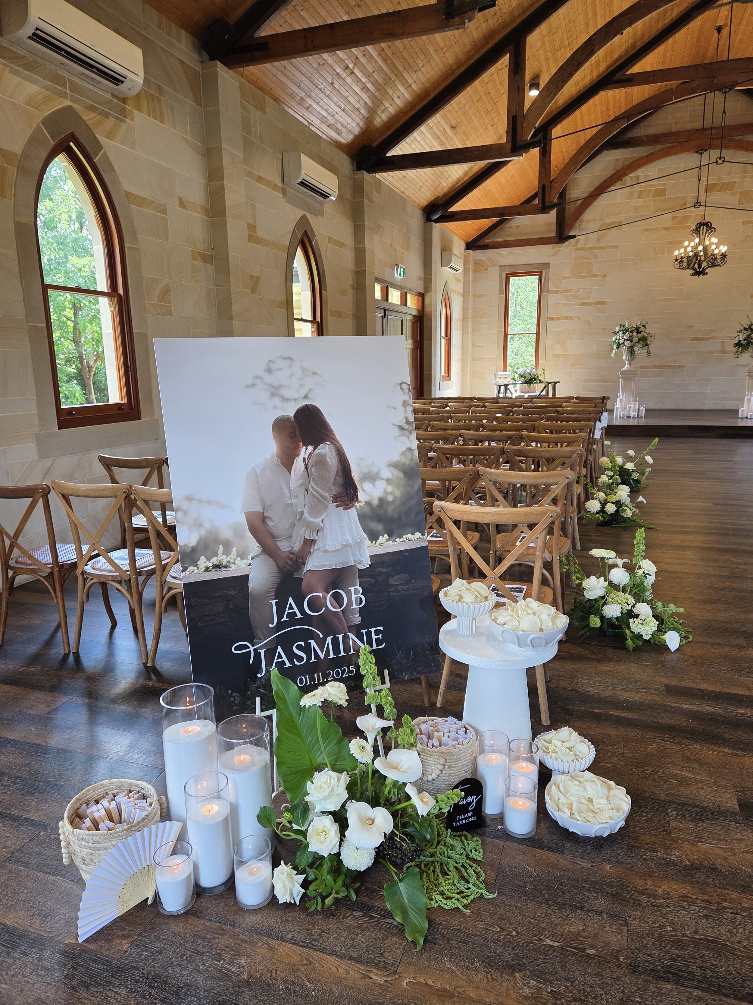 Wedding reception decor featuring a large poster of the couple Jacob and Jasmine with their wedding date, surrounded by candles, white flowers, and decorative items in a wooden hall with arched windows.