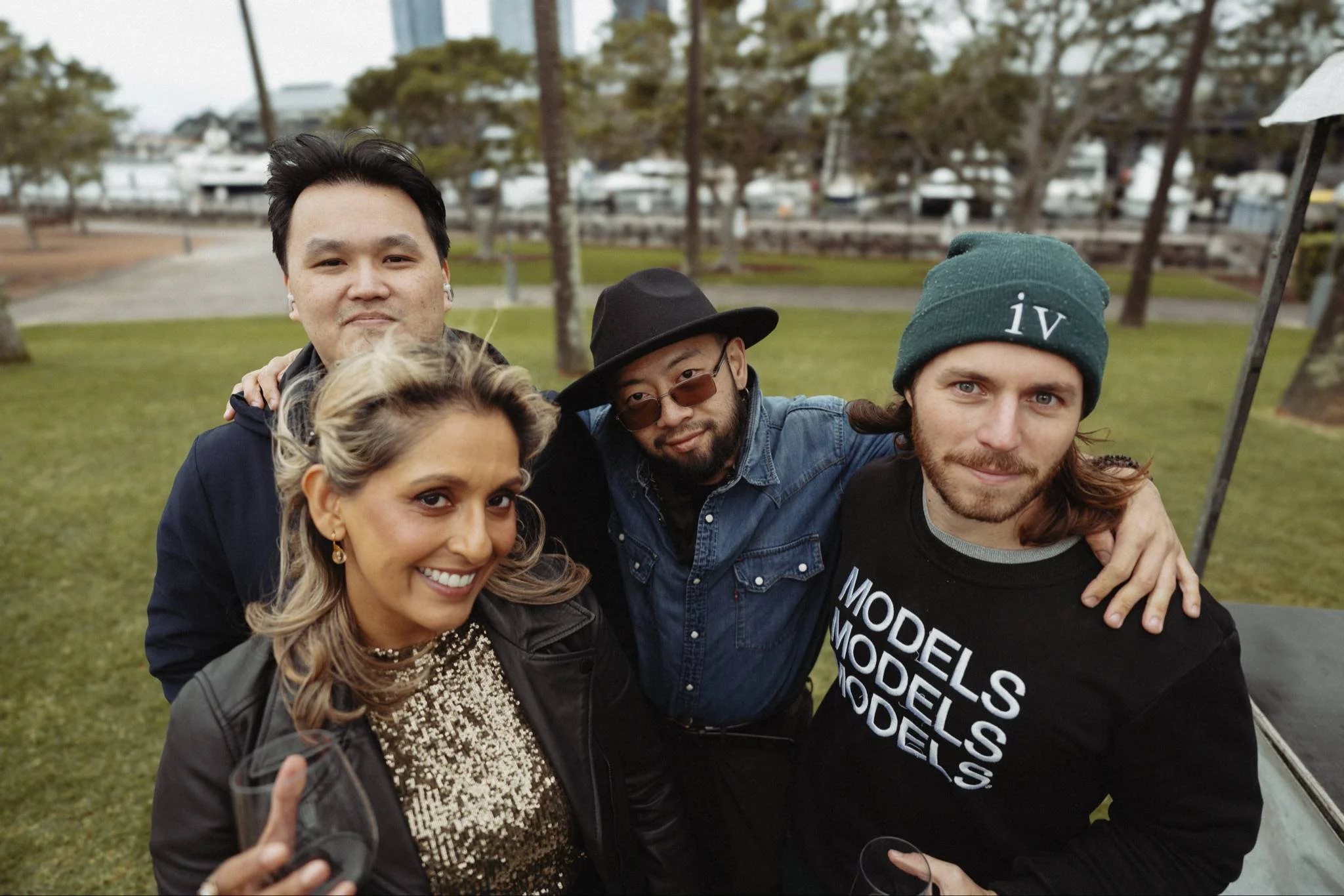 Group of four friends smiling and posing outdoors. Background includes grass, trees, and a marina with boats.