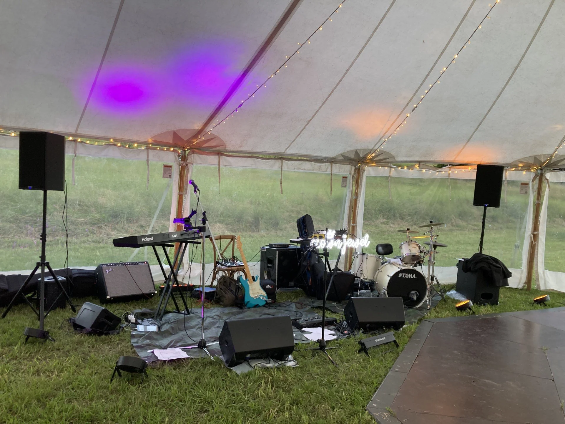 Music stage setup under a tent with instruments including a drum set, keyboard, electric guitar, and amplifier, along with speakers and microphones, decorated with string lights and a neon sign reading 'being javel'
