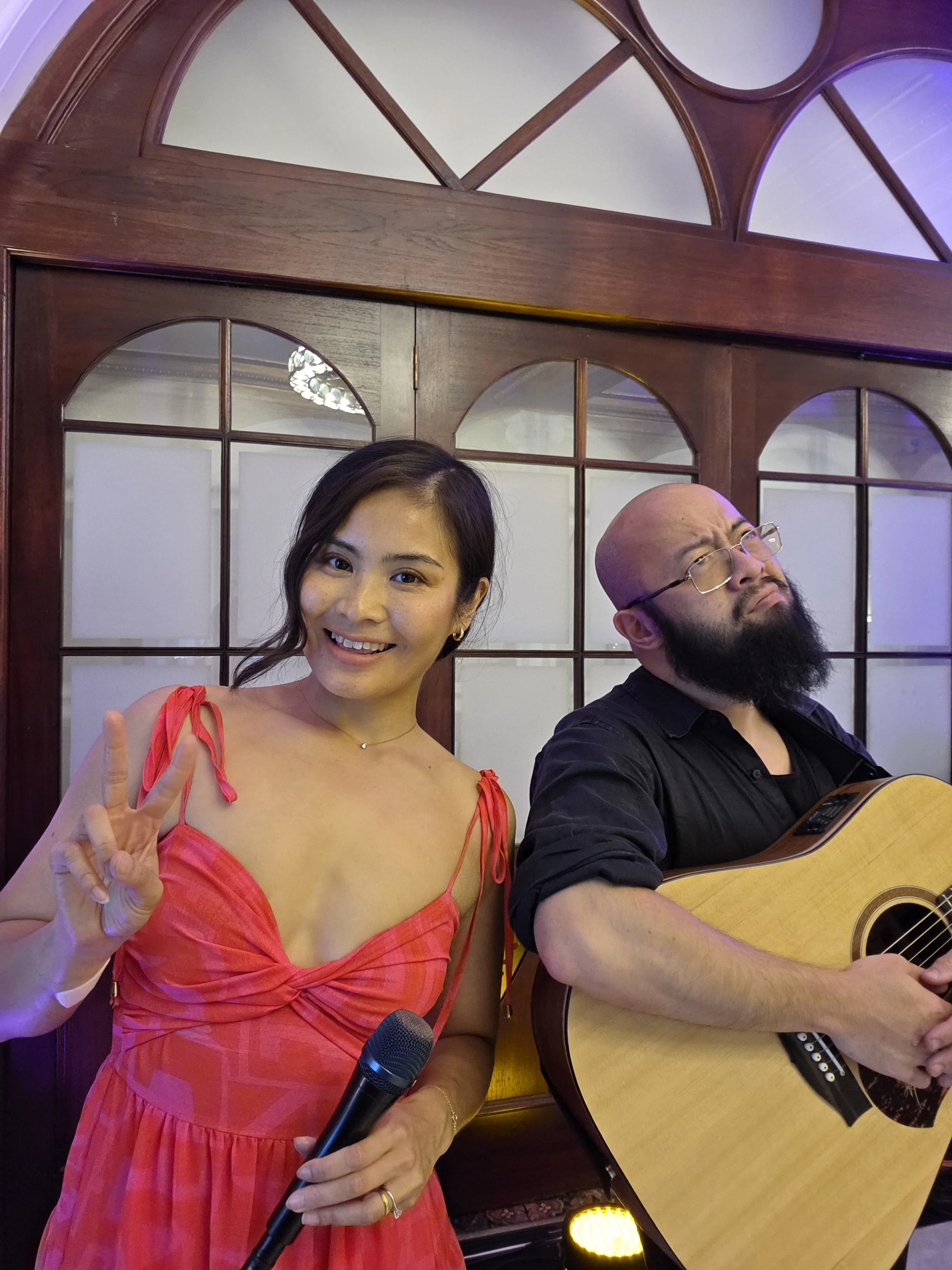 A woman in a red dress holding a microphone making a peace sign next to a man with a beard playing an acoustic guitar.