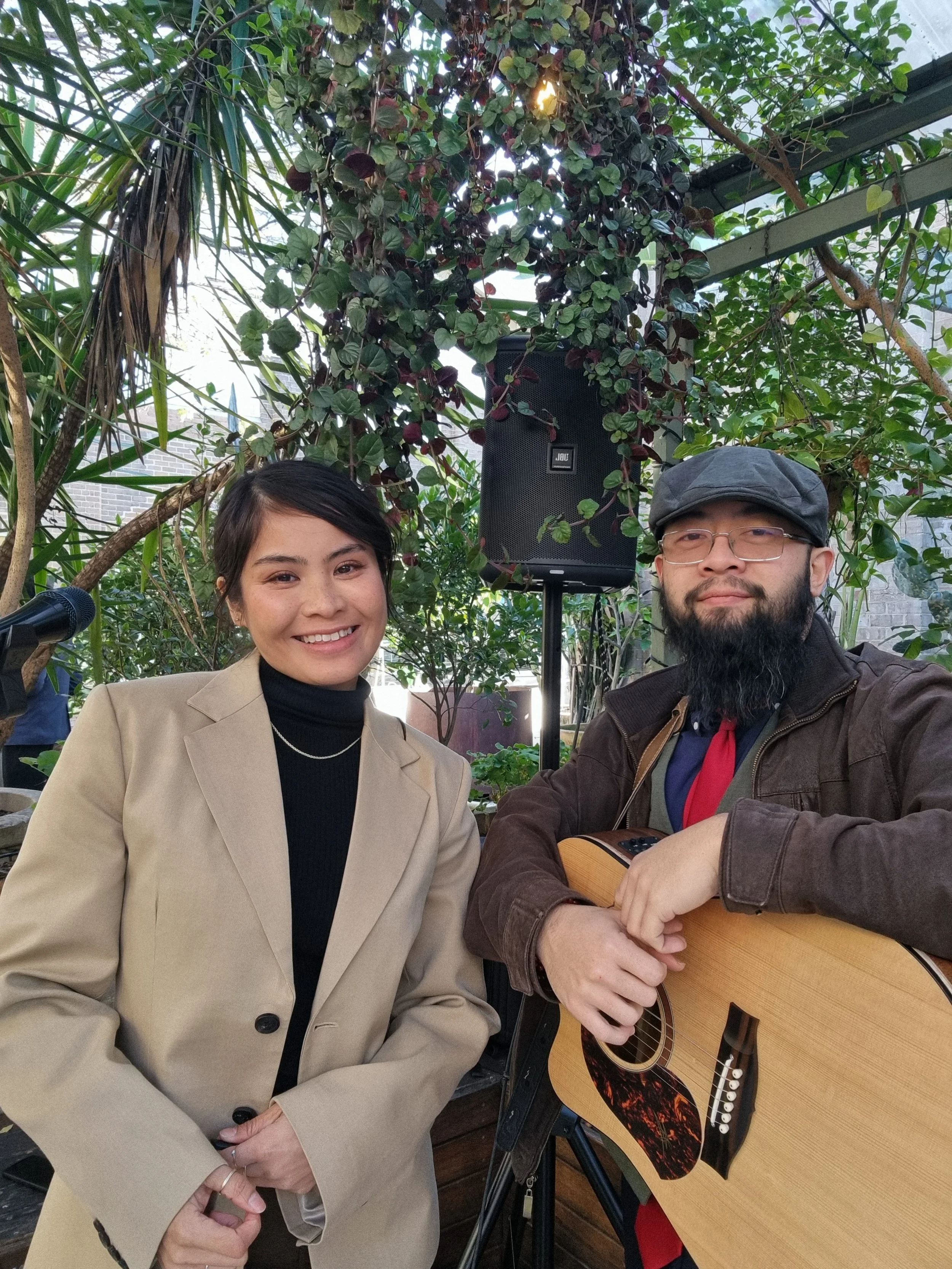 A woman and a man with a guitar posing together in a greenhouse or garden setting, with greenery and plants in the background.