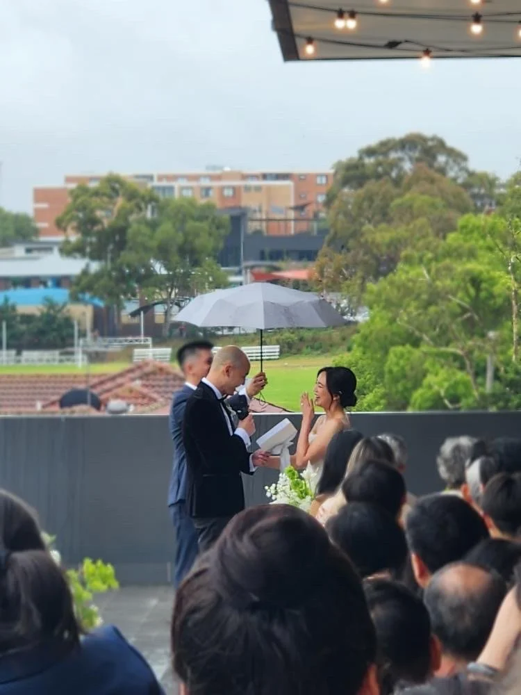 A wedding ceremony outdoors with a bride and groom under an umbrella, surrounded by seated guests, and a cityscape with trees and buildings in the background.