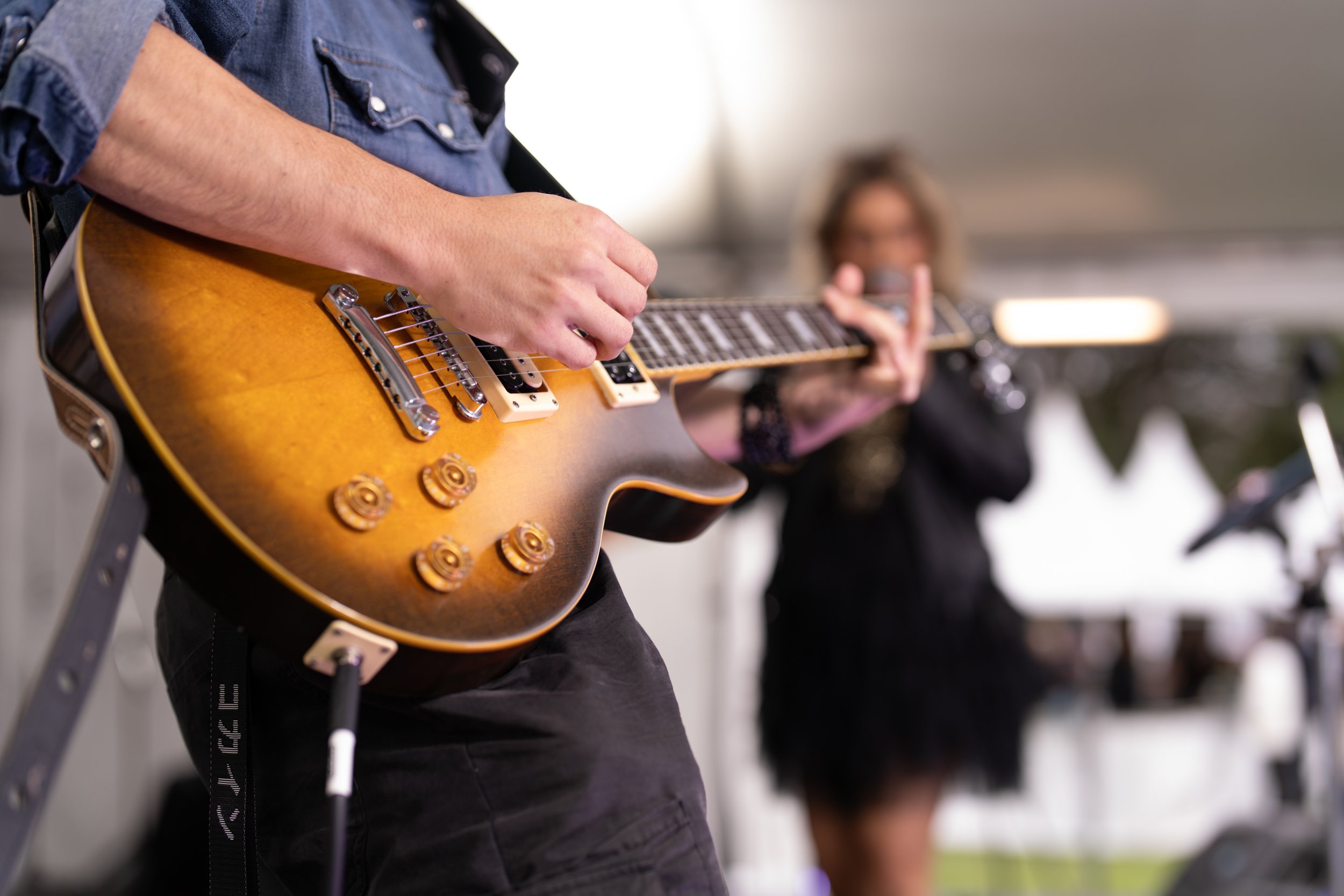 Close-up of a person playing an orange and black electric guitar, with a woman in the background holding a microphone in what appears to be a performance setting.