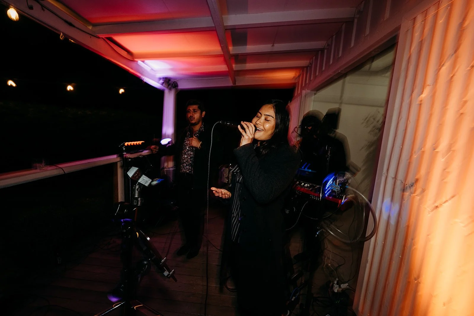 A woman singing into a microphone on a small outdoor stage at night, with two men behind her, one with DJ equipment, under colorful lighting.
