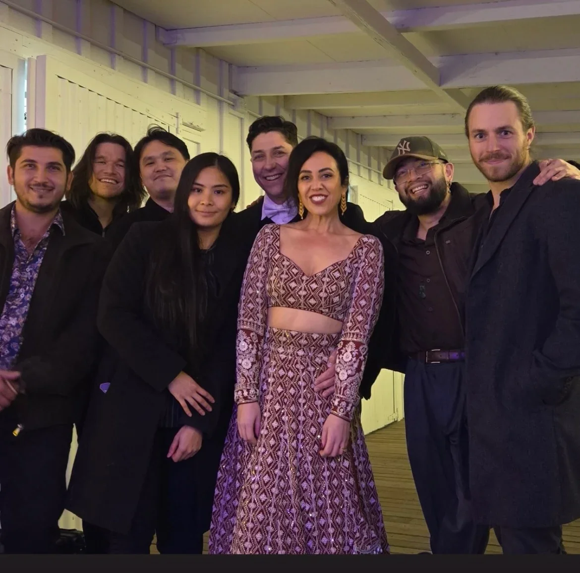 Group of eight people, five men and three women, posing indoors at a social event, with some dressed in formal wear and casual attire.