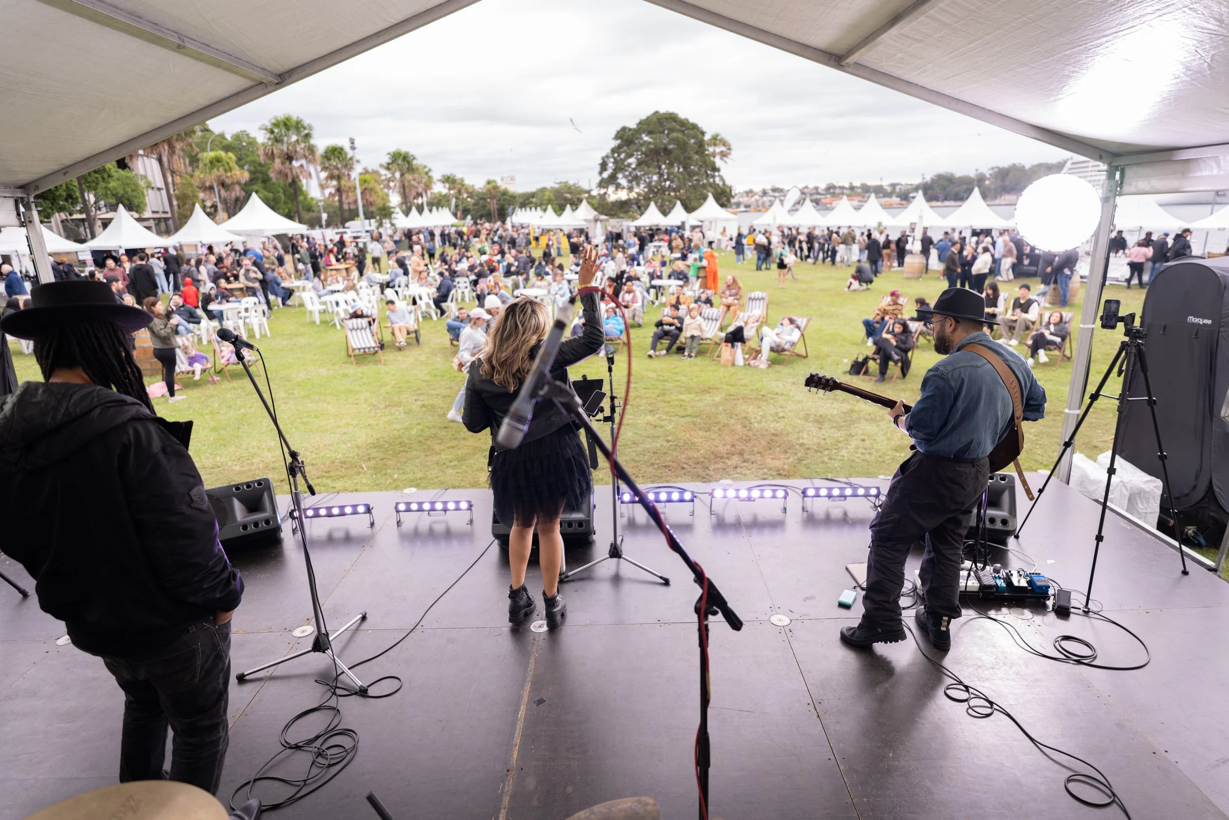 Band performing on stage at outdoor festival with audience seated on chairs and standing on grassy field, white tents in background, daytime with overcast sky.