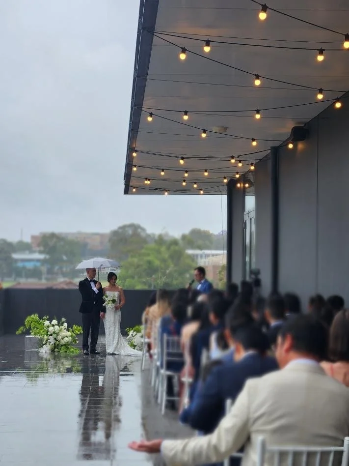 A wedding ceremony taking place on an outdoor terrace on a rainy day, with the bride holding a bouquet and standing next to the groom under an umbrella, with guests seated under string lights.