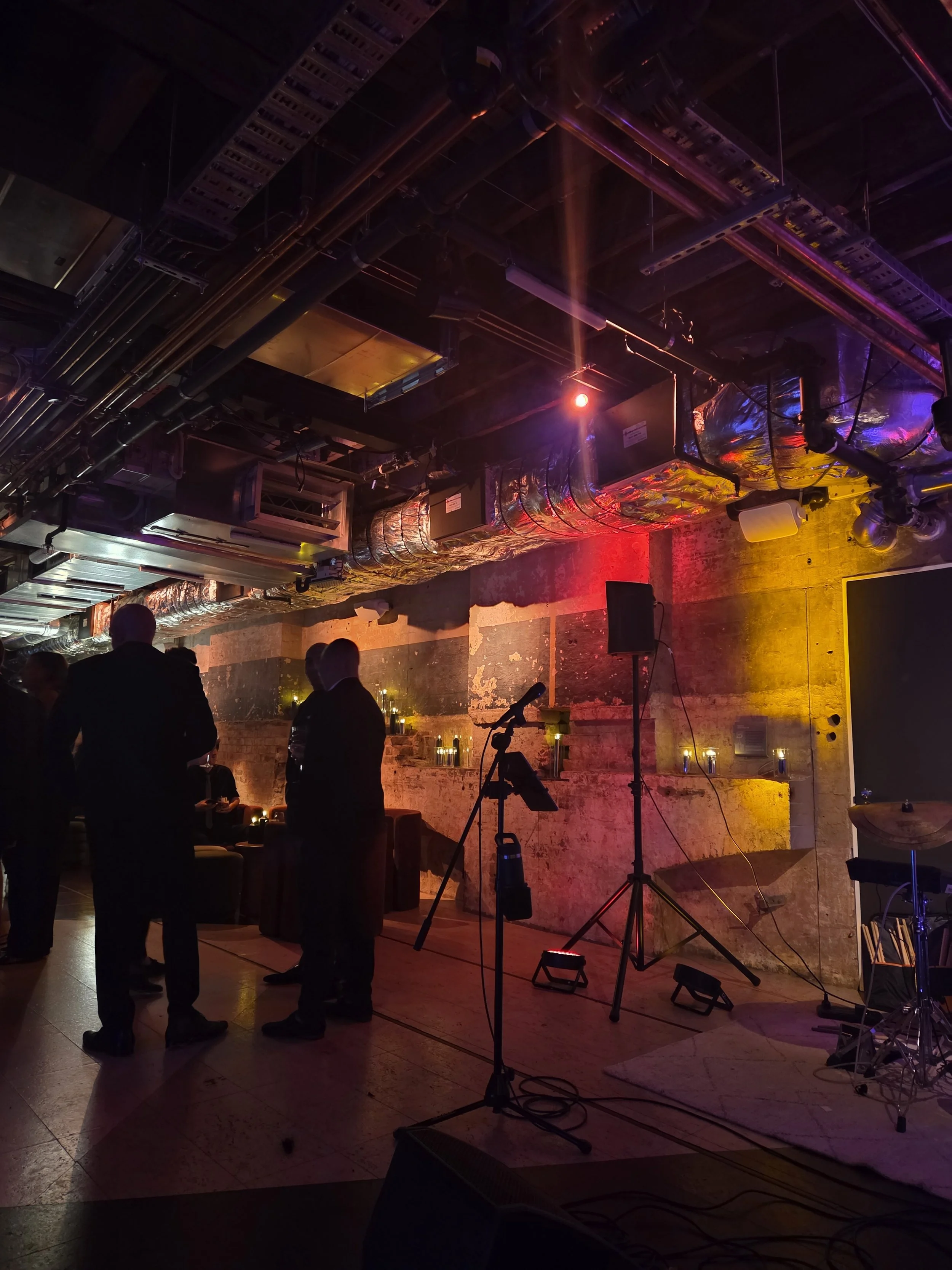A dimly lit indoor venue with a stage set up for a performance, including microphones, a speaker, and drums, with abstract wall art and colorful lighting.