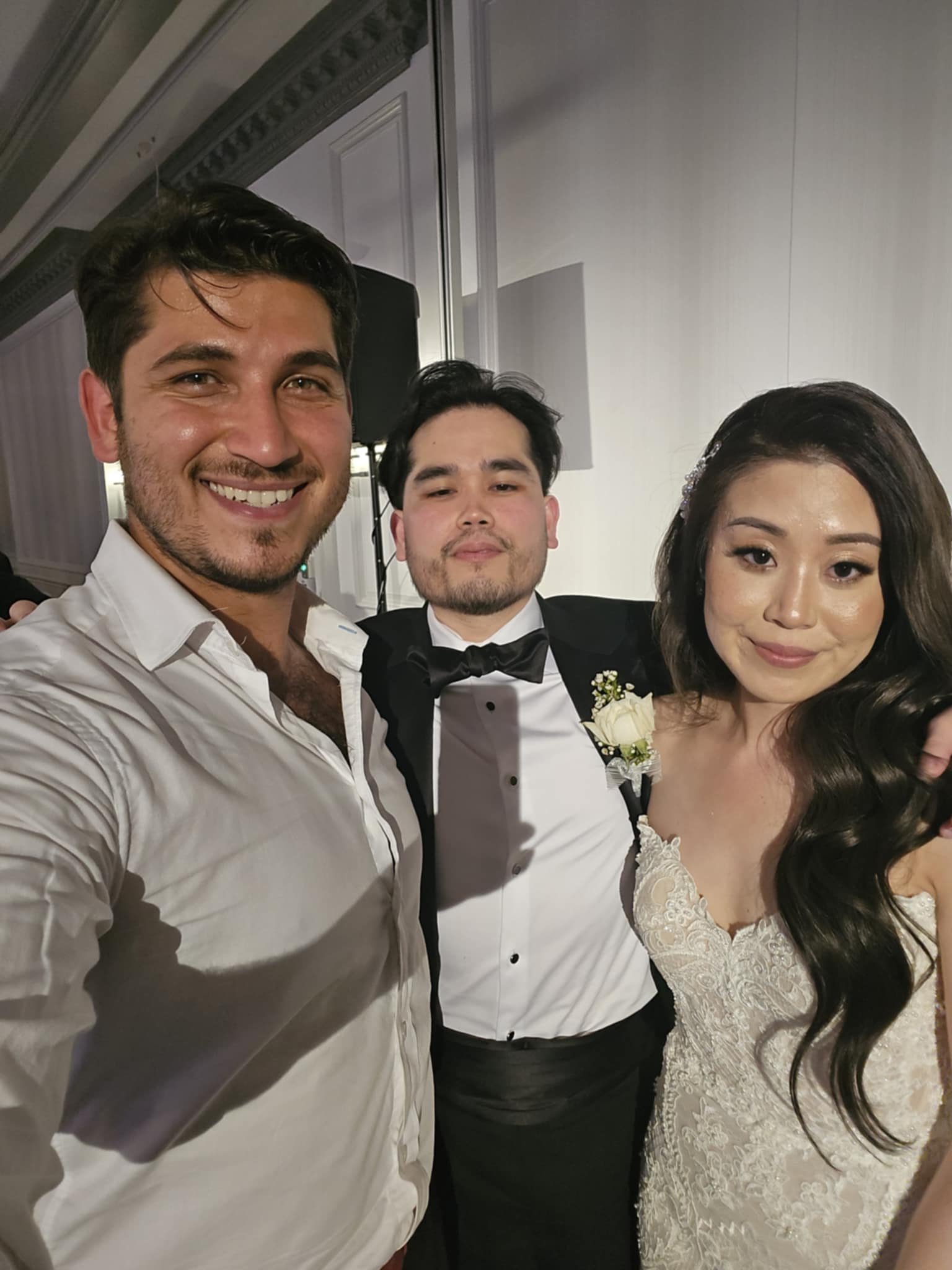 Three people, a man and a woman in wedding attire and another man in formal tuxedo, taking a close-up selfie at a wedding reception.