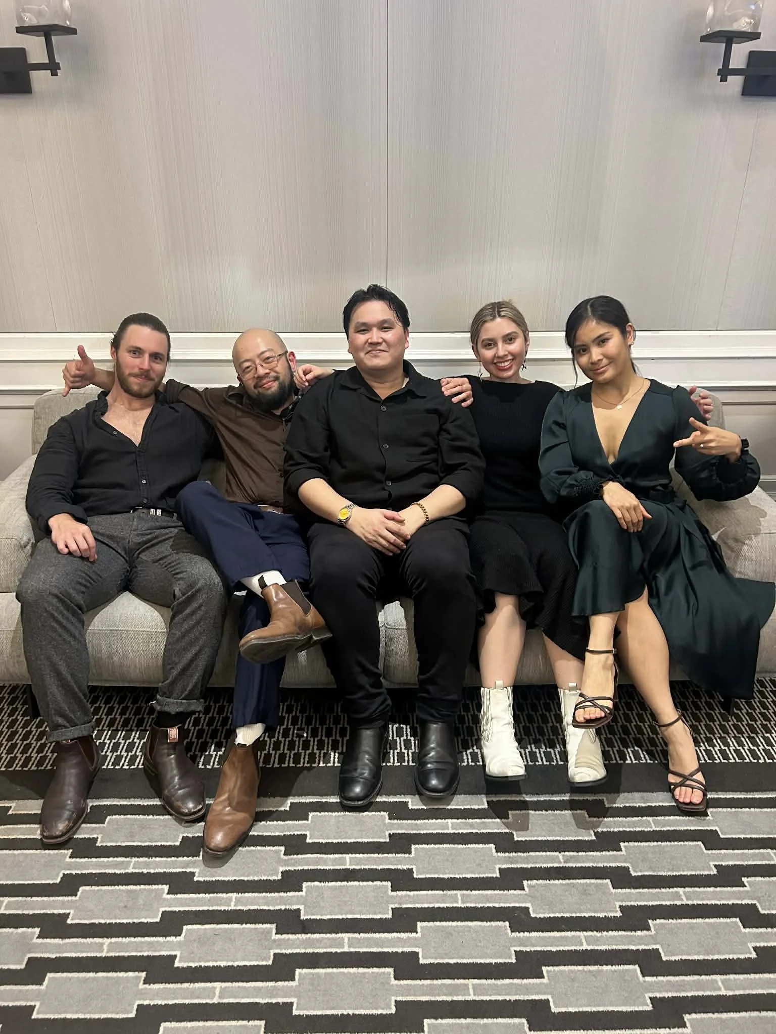 Group of five people sitting on a light-colored sofa in formal attire, smiling for the photo in a room with a patterned carpet and a plain wall behind them.