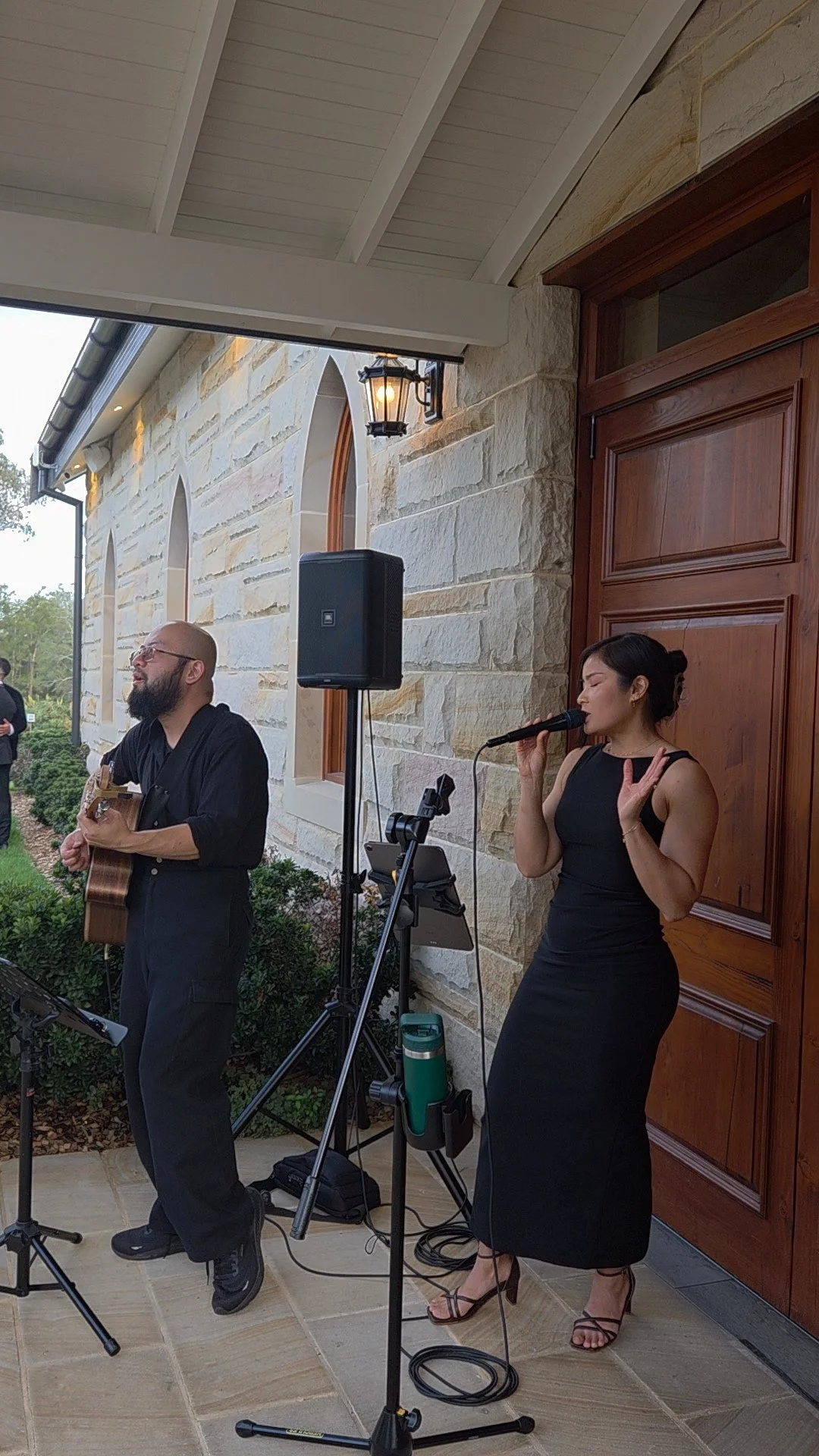 A man playing guitar and a woman singing into a microphone during an outdoor performance in front of a stone building with wooden door and window.