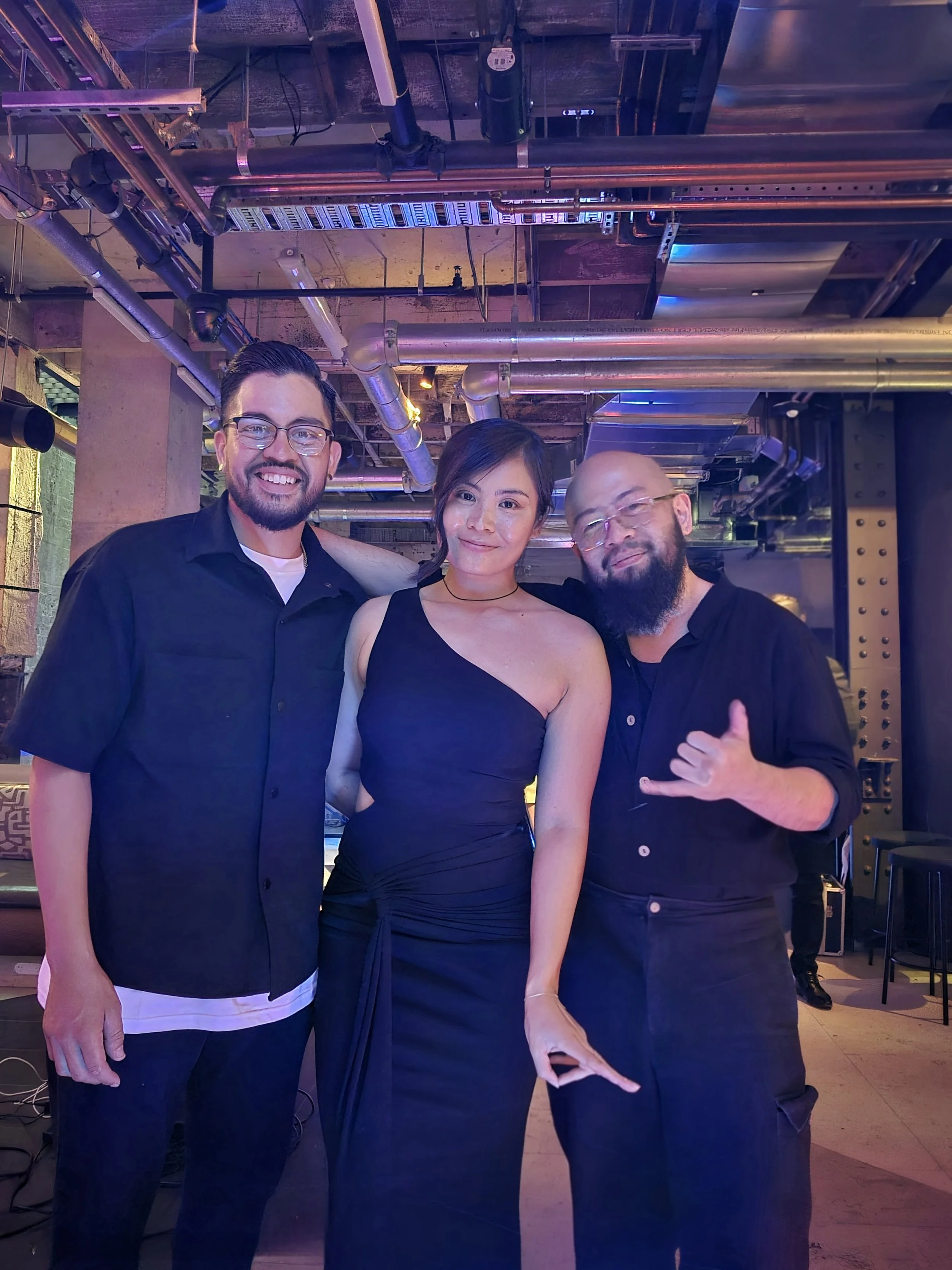 Three people smiling and posing together in a dimly-lit, industrial-style space with exposed pipes and metal structures.