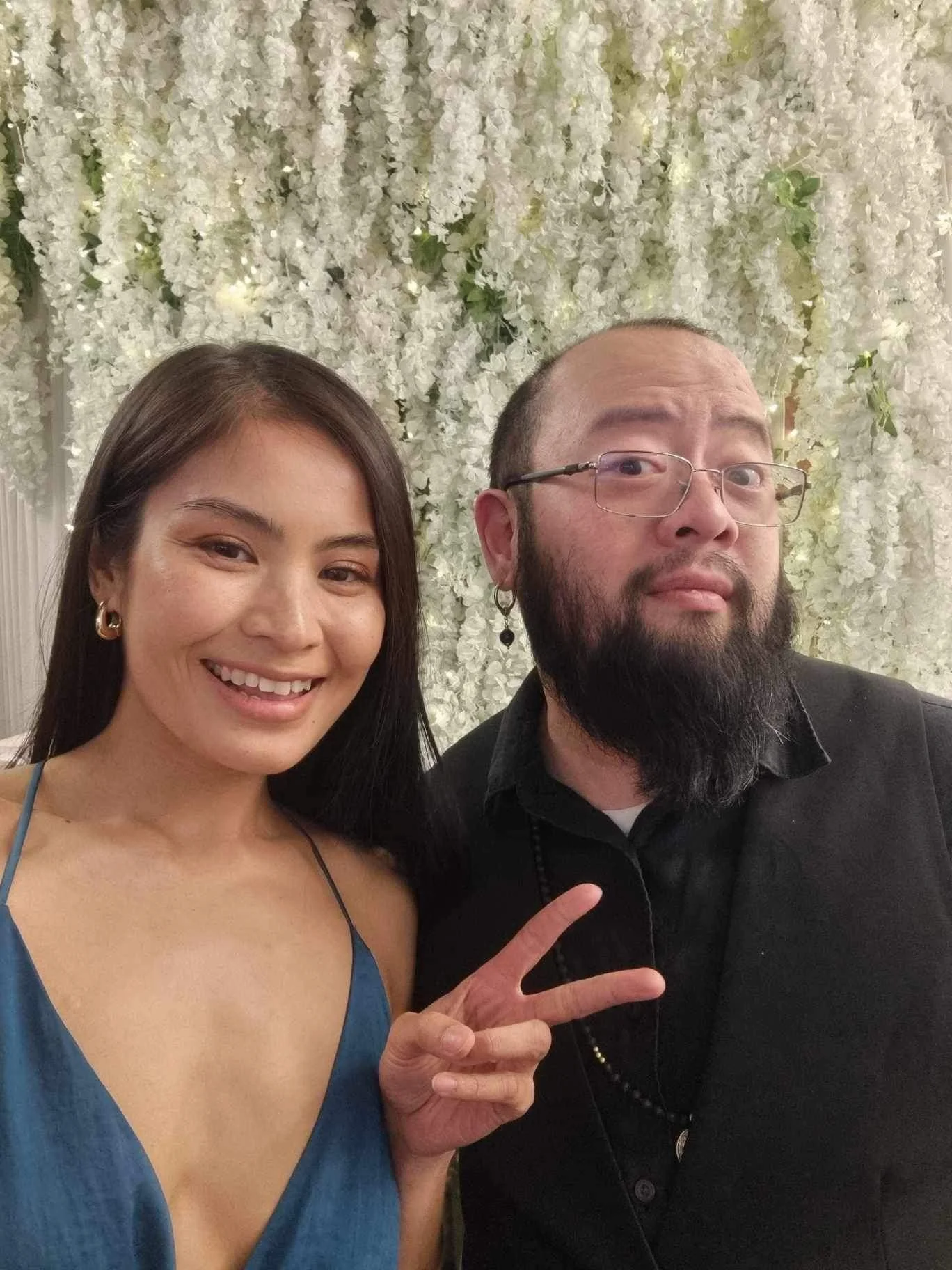 A woman smiling and making a peace sign with her hand next to a man with glasses and a beard, standing in front of a white floral backdrop.