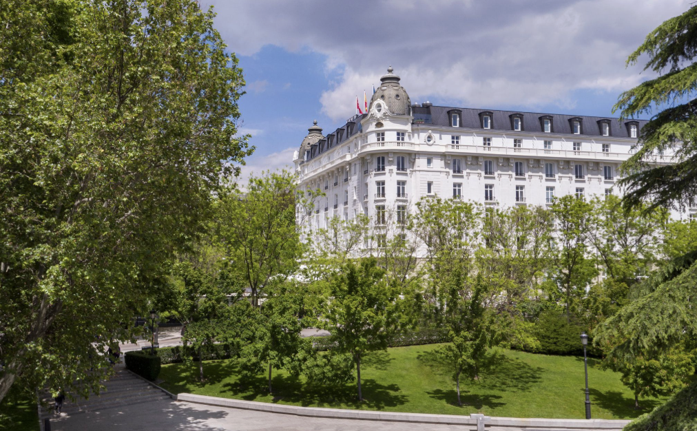 A large white hotel building with dark roof accents, surrounded by green trees and a grassy park area, under a partly cloudy sky.
