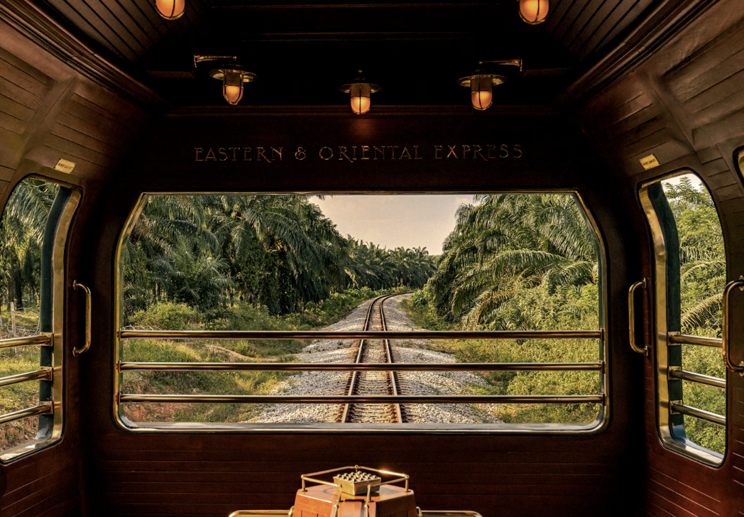 View from inside a train labeled 'Eastern & Oriental Express' looking out the window at train tracks and lush green palm trees.