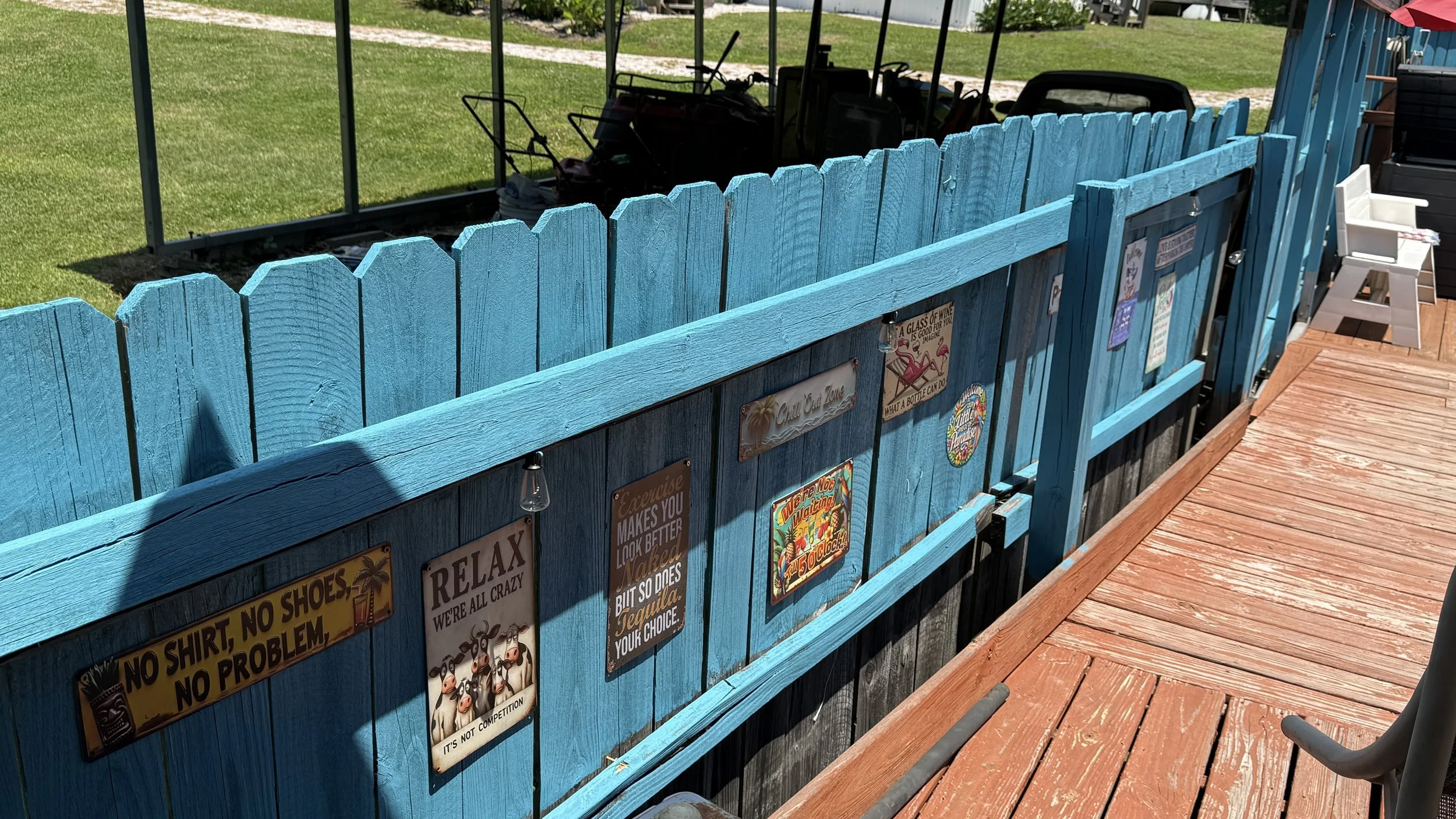 Blue wooden fence decorated with various humorous signs, next to a wooden deck and a black metal railing. In the background, a grassy yard and outdoor equipment are visible.
