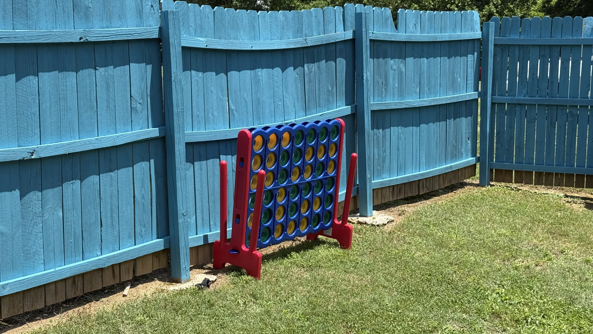 A blue wooden fence surrounds a grassy yard. In front of the fence, there is a large Connect Four game with a red and blue frame and yellow, green, and blue discs, with some discs already stacked in the game.