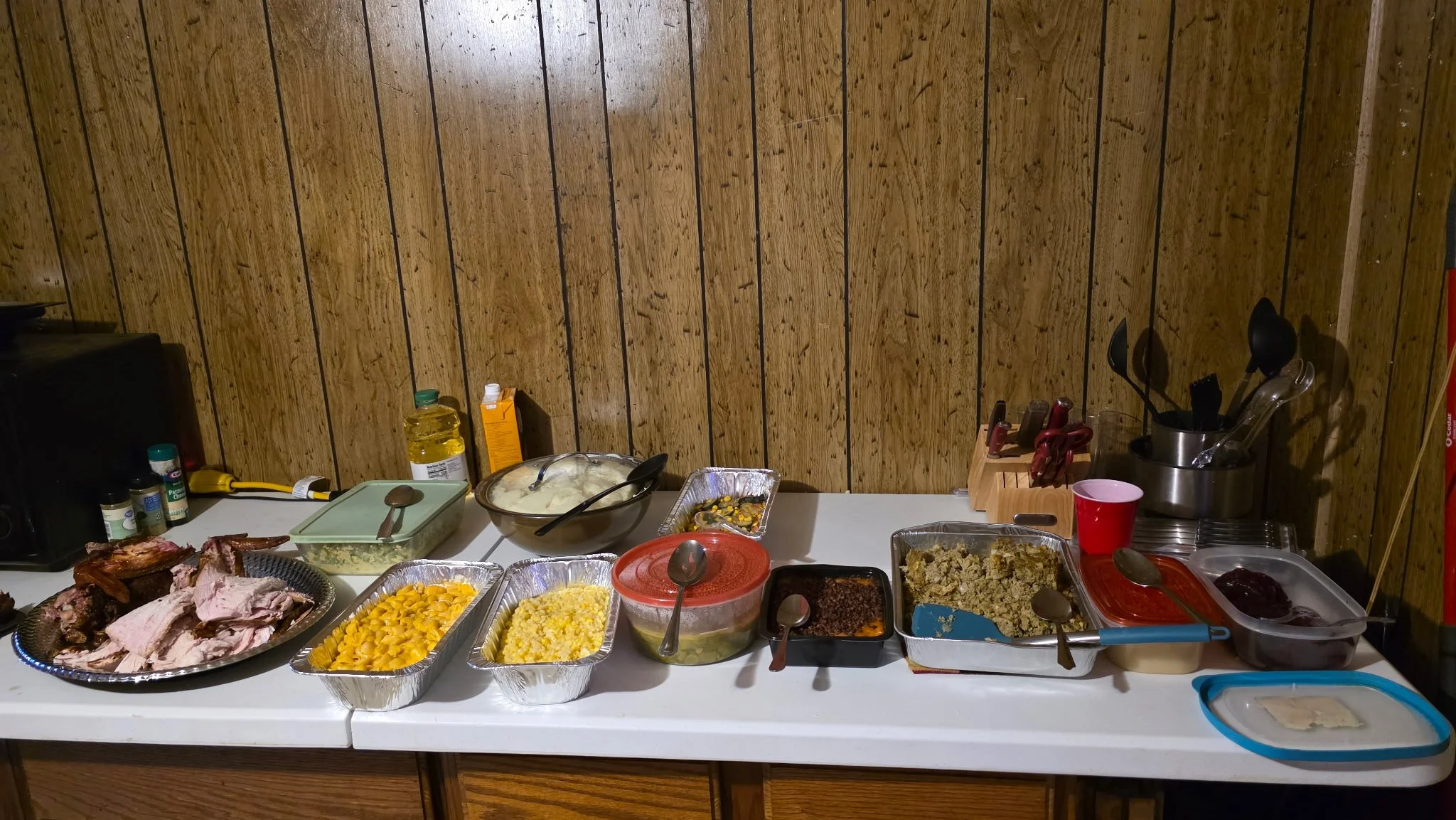 A table with various dishes of food, including meats, macaroni and cheese, corn, and other side dishes, set against a wood-paneled wall background.