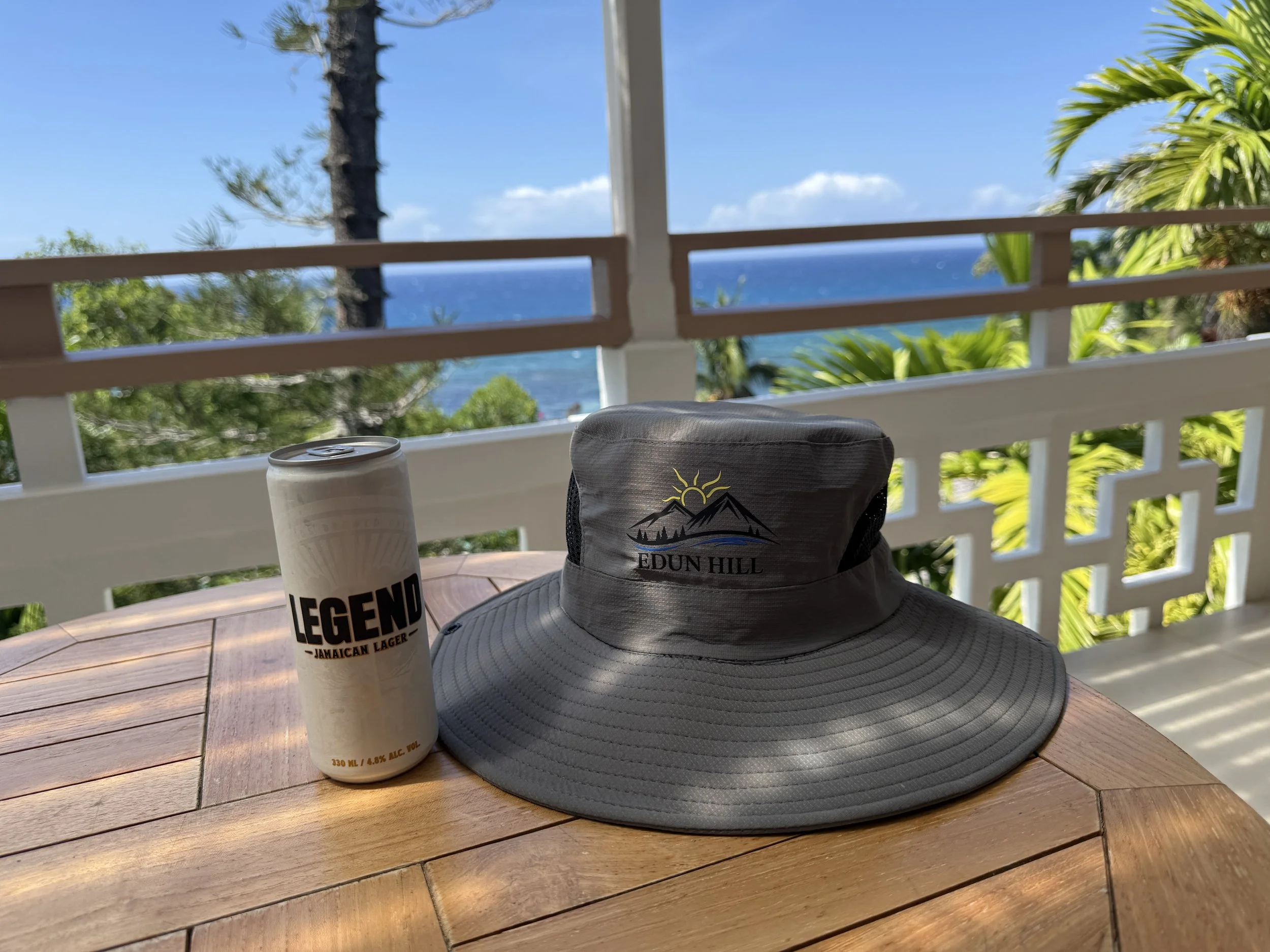 A wooden table with a gray wide-brimmed hat, a can of Legend Jamaican Lager, and a gray water bottle with a sun and mountain logo, set on a porch overlooking trees, the ocean, and a clear blue sky.