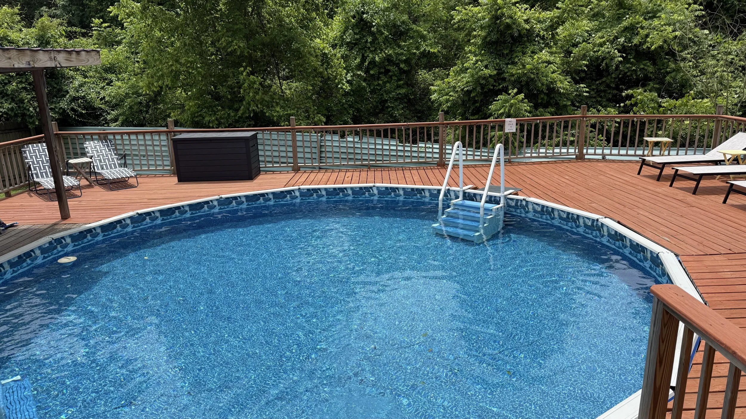 Round swimming pool with blue water and steps, surrounded by a wooden deck with lounge chairs and a black storage box, backed by green trees.