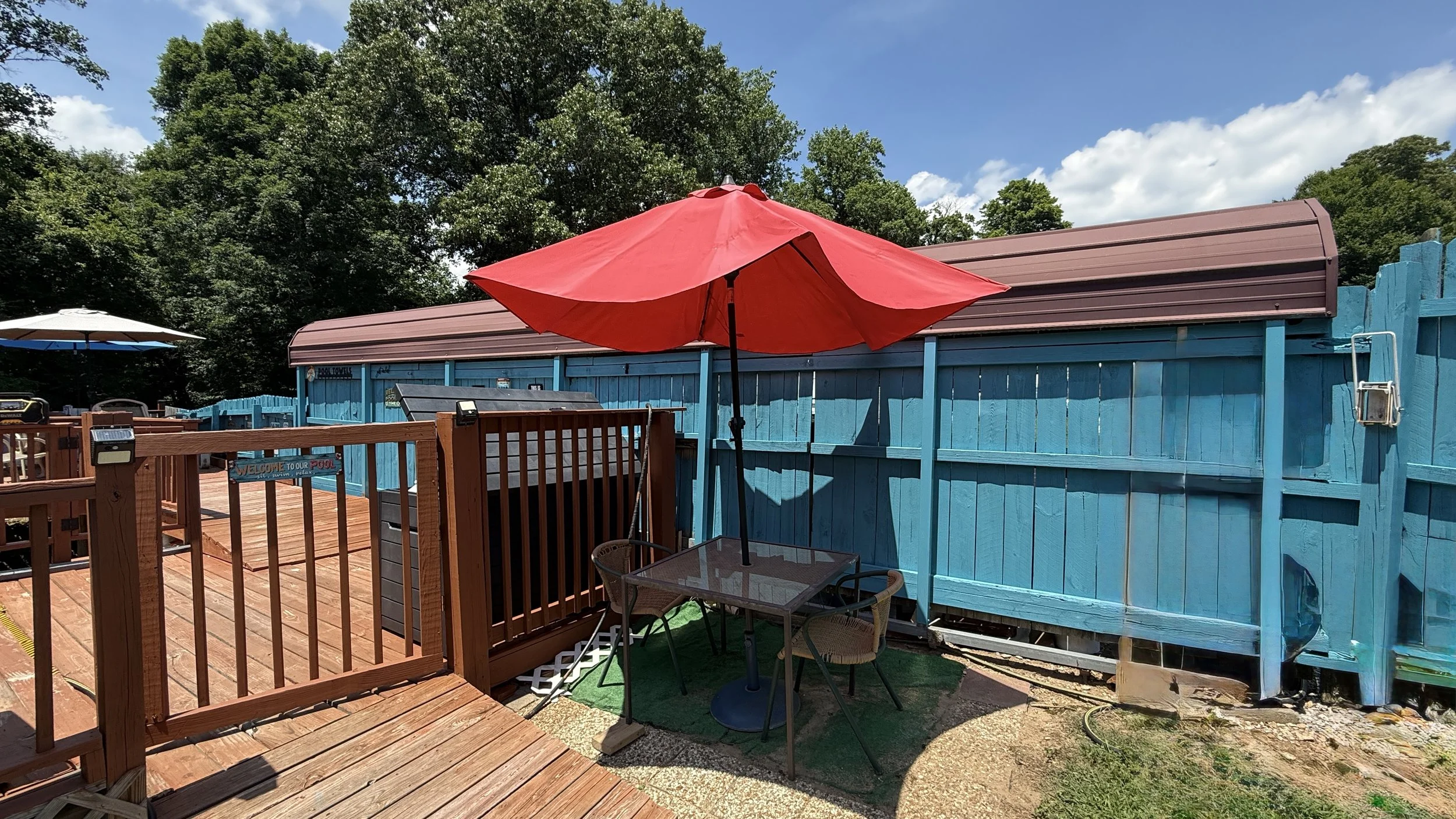 Backyard with a wooden deck, a red patio umbrella, and a blue wooden fence. There are trees and a partly cloudy sky in the background.