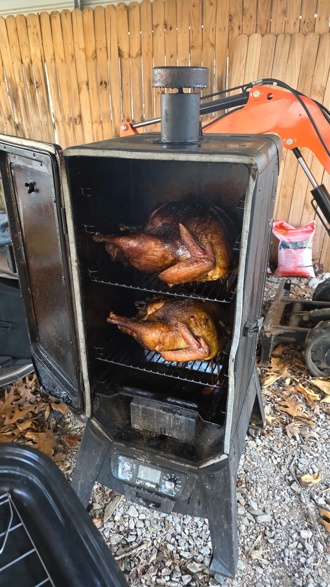 Two whole turkeys roasting in a black electric smoker with an open door, set outdoors on ground covered with fallen leaves and a wooden fence in the background.