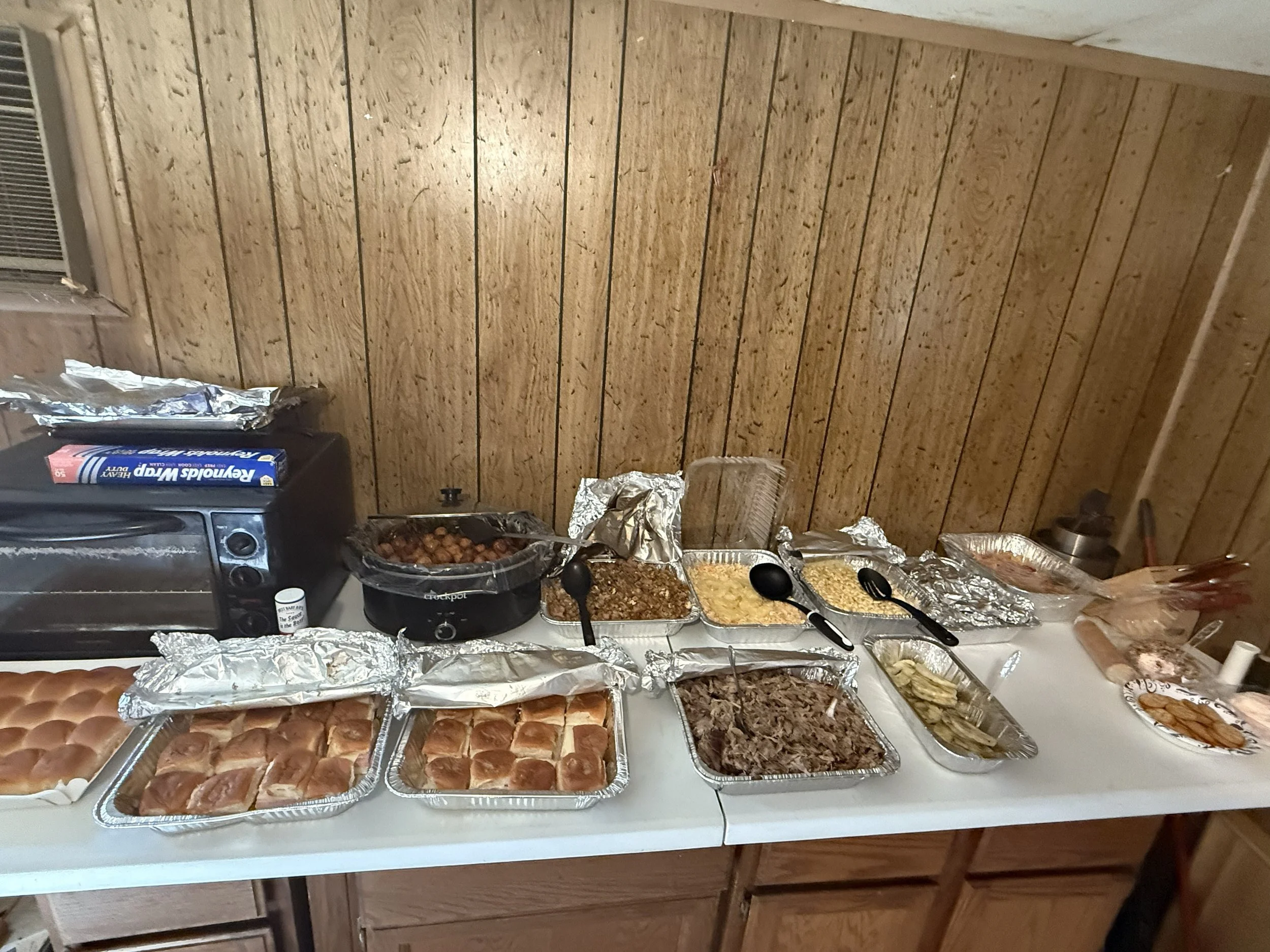 A buffet table filled with various dishes, including trays of baked foods, a crockpot with meatballs, and aluminum foil-covered containers, set against a wooden-paneled wall.