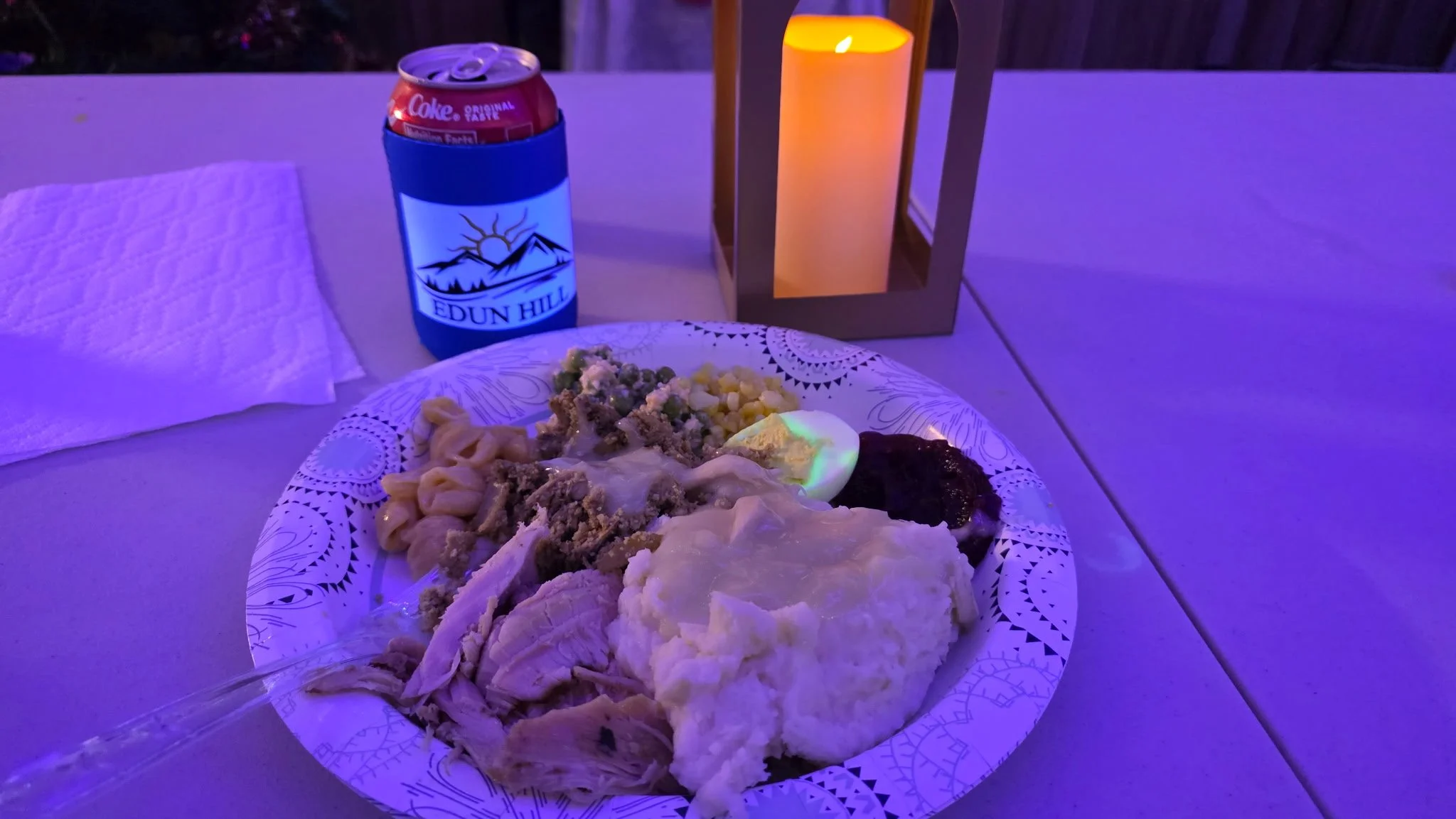 Plastic plate with assorted foods including mashed potatoes, vegetables, and meat, on a white table. In the background, there is a blue can holder with a soda can, a paper towel, a candle inside a decorative lantern, and purple lighting.