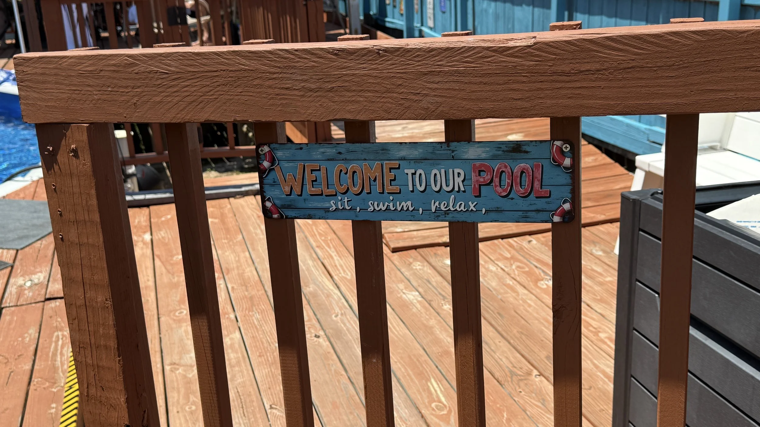 Wooden deck with a blue sign that reads "Welcome to our Pool sit, swim, relax" attached to a brown railing. The background shows parts of a pool and other wooden structures.