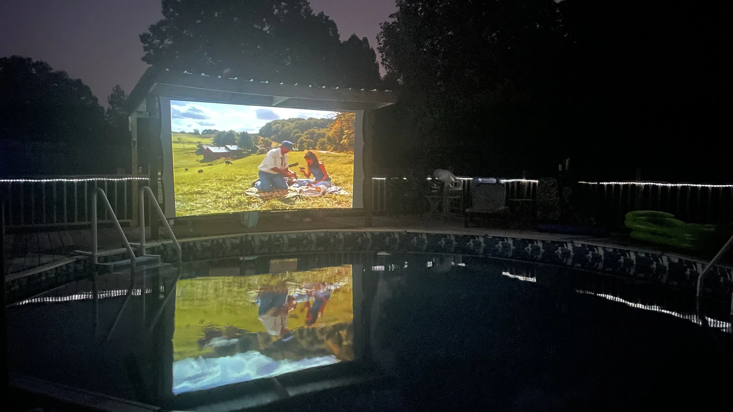 An outdoor swimming pool at night with a large flat-screen outdoor movie projector displaying an image of two people having a picnic on grass with trees and a blue sky in the background.
